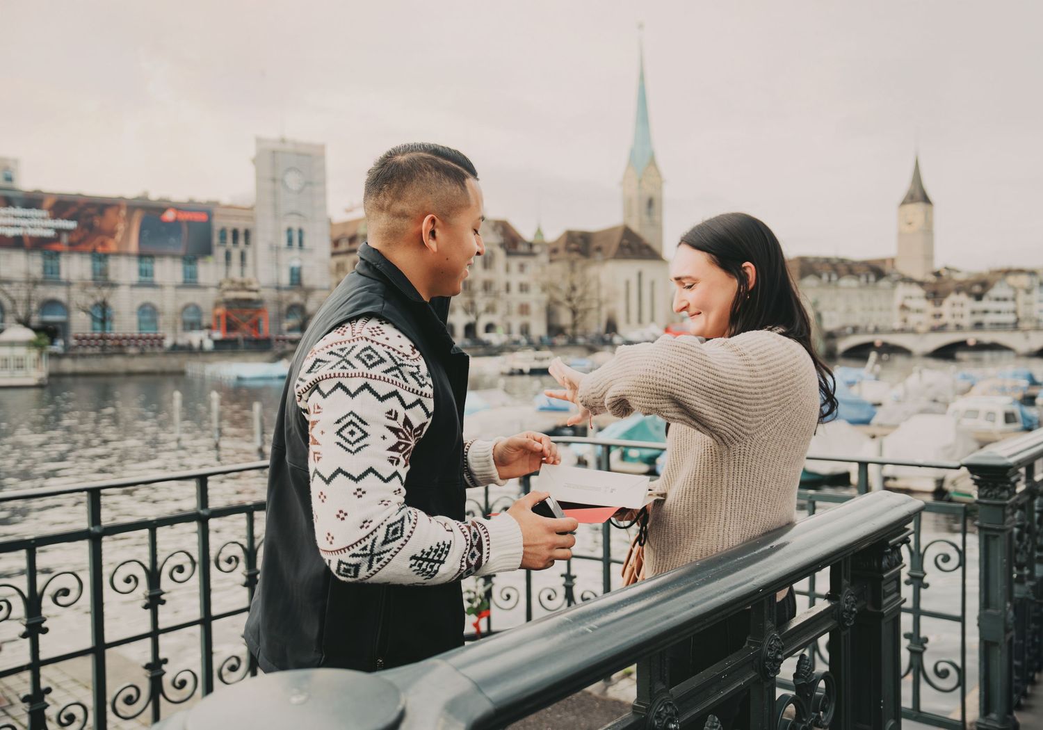 Couple exchanging rings on Zurich waterfront bridge with historic buildings in background.