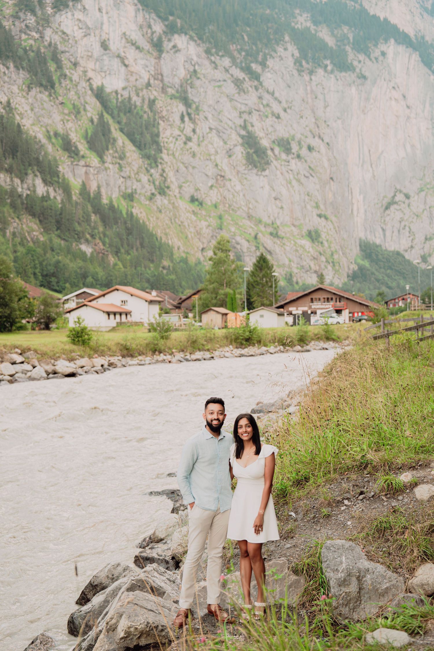 Couple standing on rocky riverbank with Swiss Alpine village and dramatic mountain cliffs in the background.