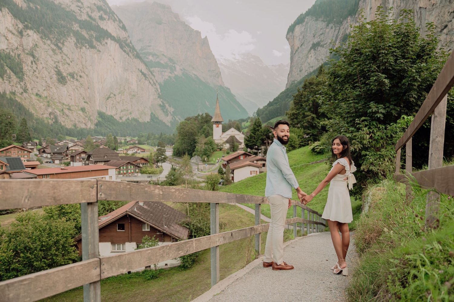 Couple holding hands on a scenic overlook in Lauterbrunnen, Switzerland, with alpine village and misty mountains behind.