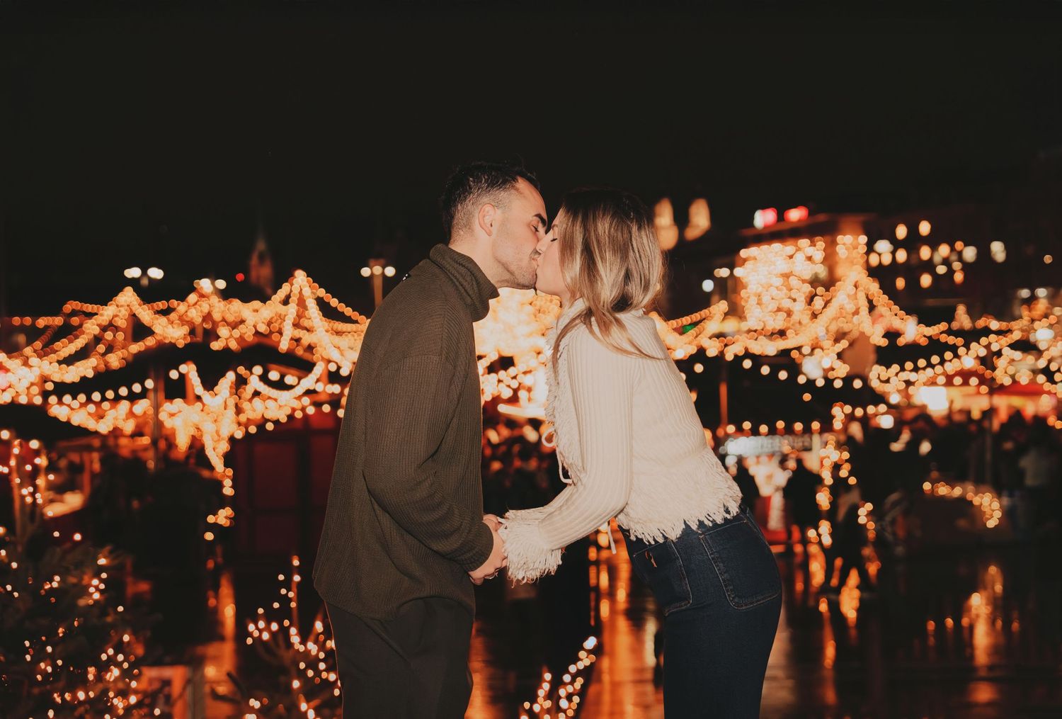 Couple sharing a kiss at night surrounded by warm glowing Christmas market lights.
