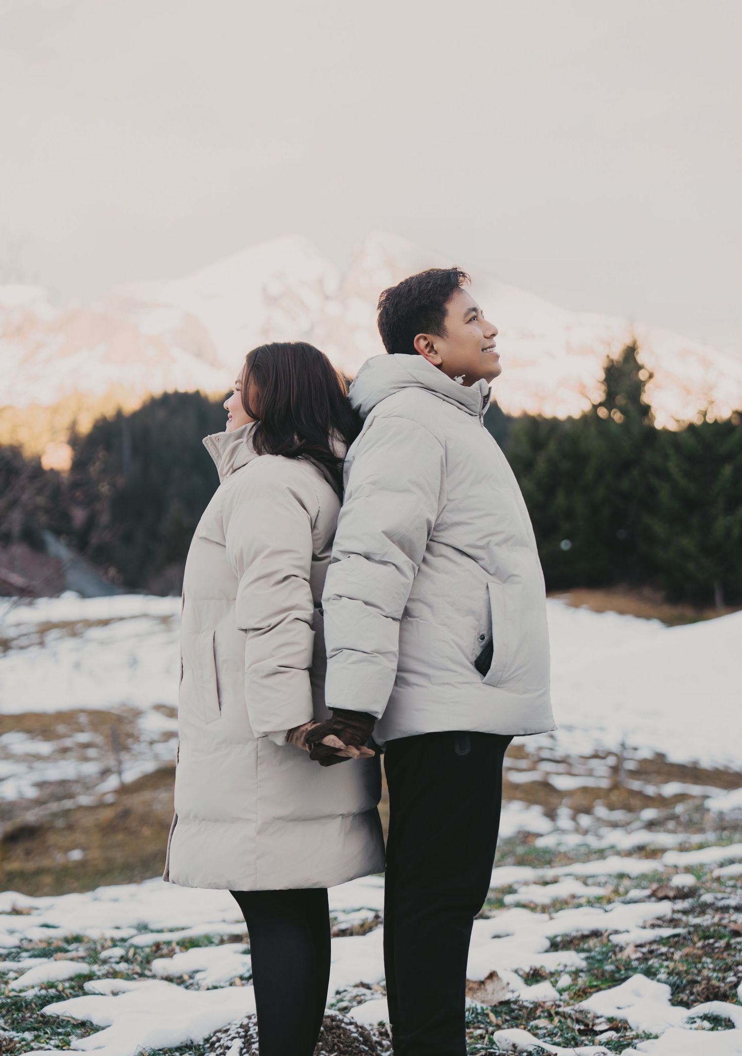 Couple standing back to back in matching grey puffer coats in snowy mountain setting.