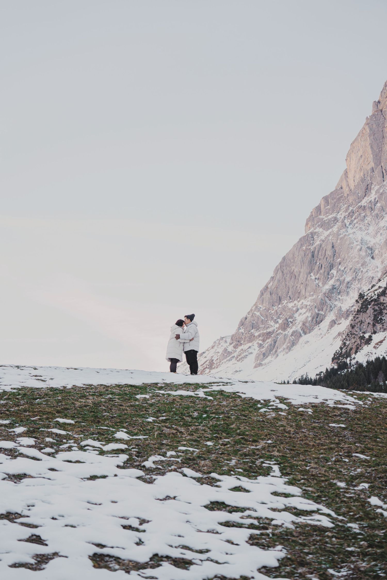 Couple embracing on snowy alpine meadow with dramatic rocky mountain peaks in winter landscape.