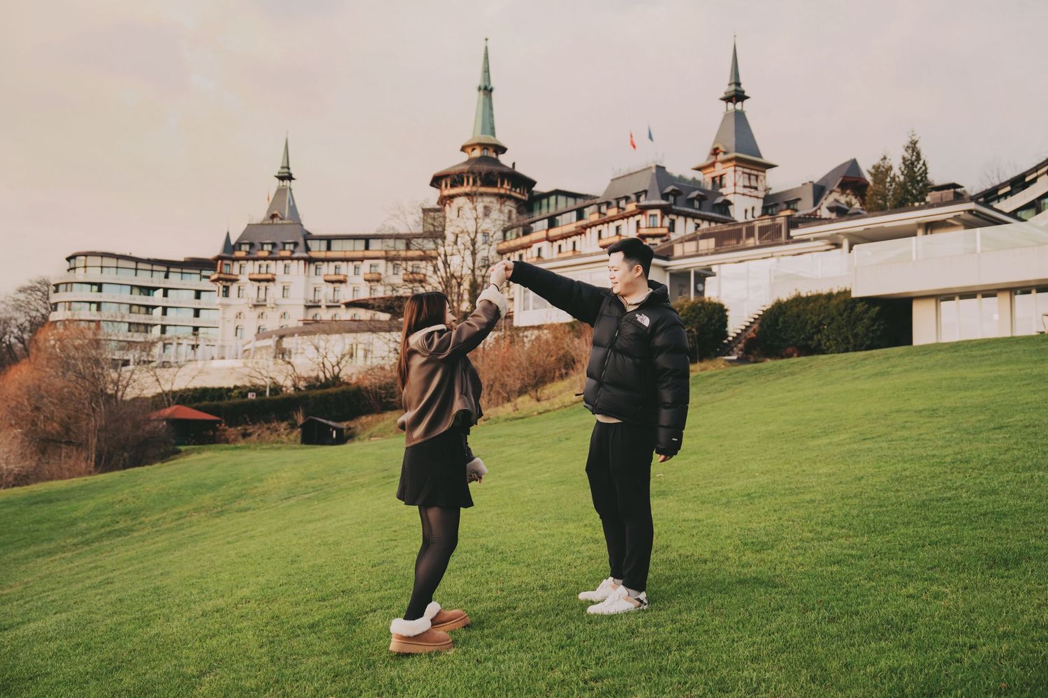 Couple playfully dancing on green lawn in front of grand European castle-style hotel.
