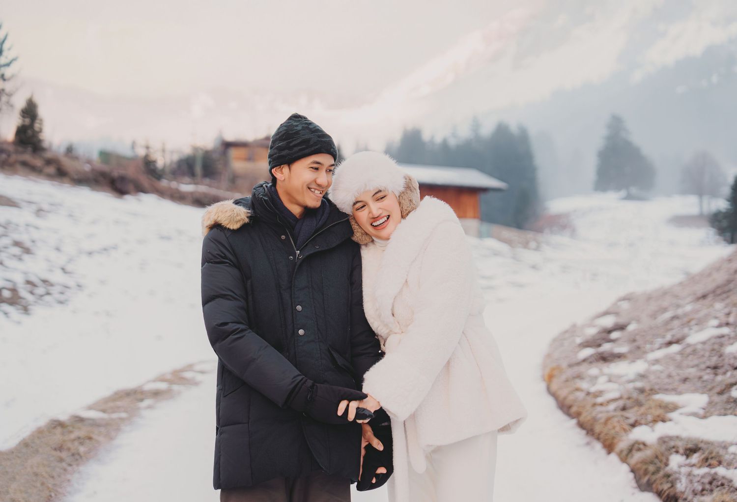Laughing couple in winter coats on snowy Alpine road with soft mountain and pine tree scenery.