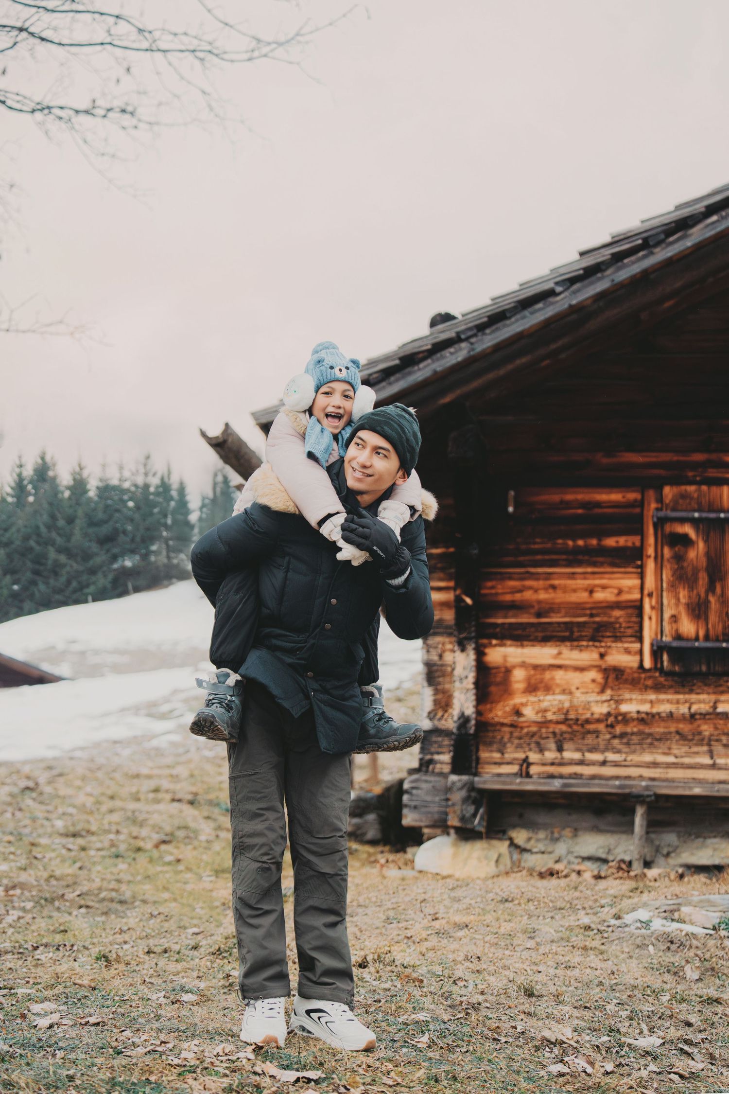 Man giving laughing child a piggyback ride outside rustic wooden cabin in snowy alpine winter.