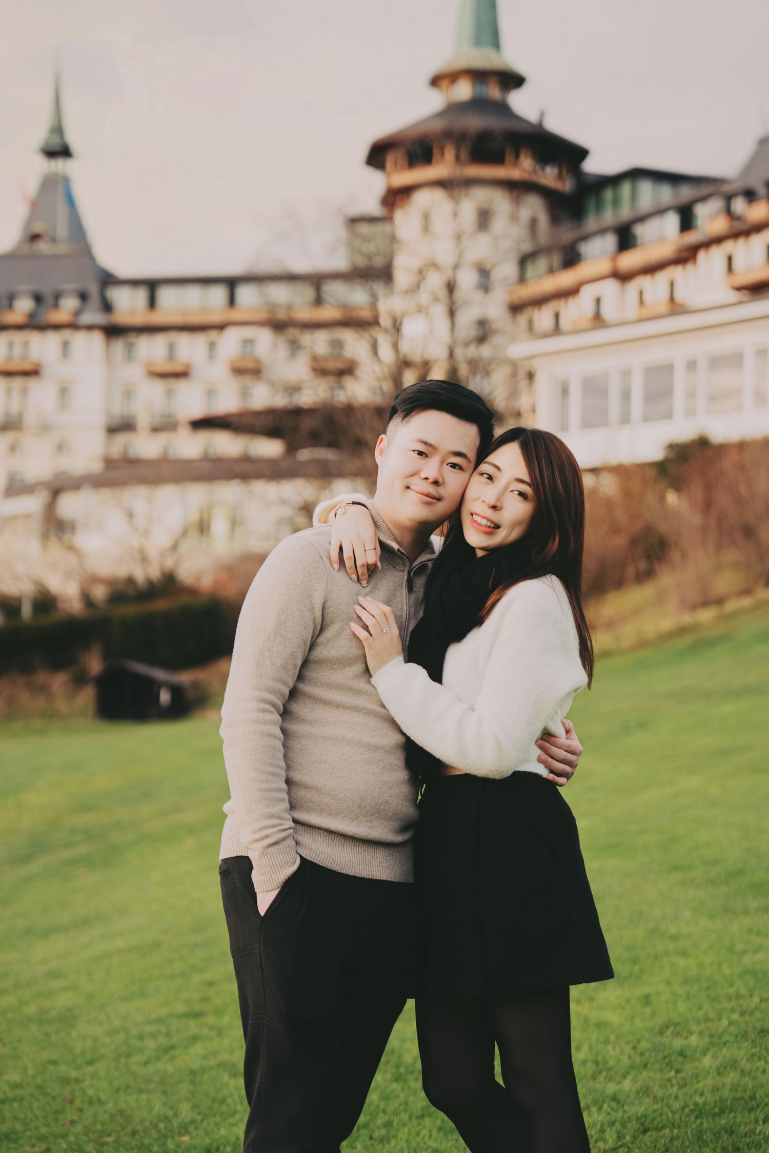 Couple hugging and smiling on a lawn with a large European luxury hotel in the background.