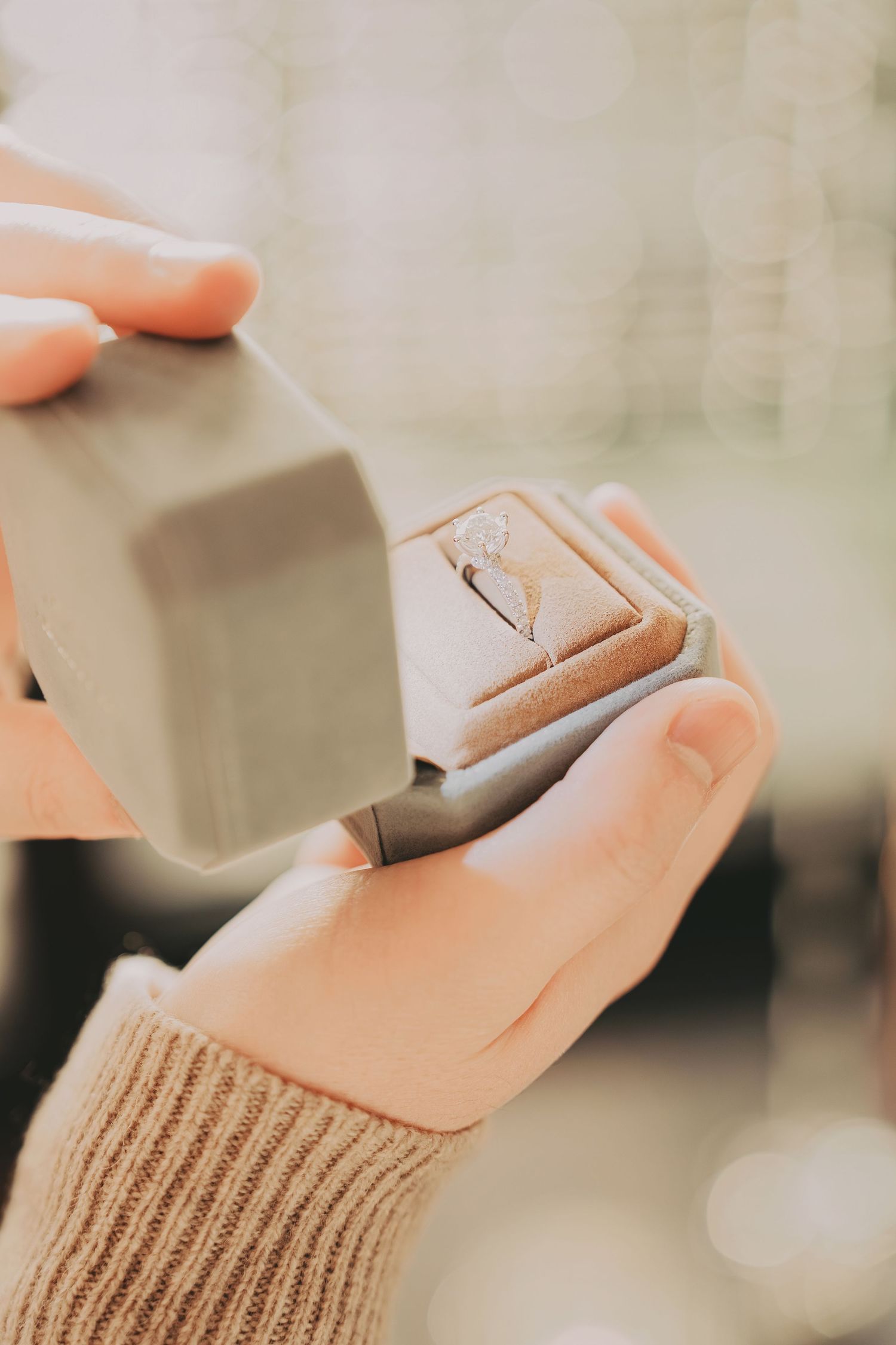 Close-up of hands holding open gray ring box with diamond engagement ring inside.