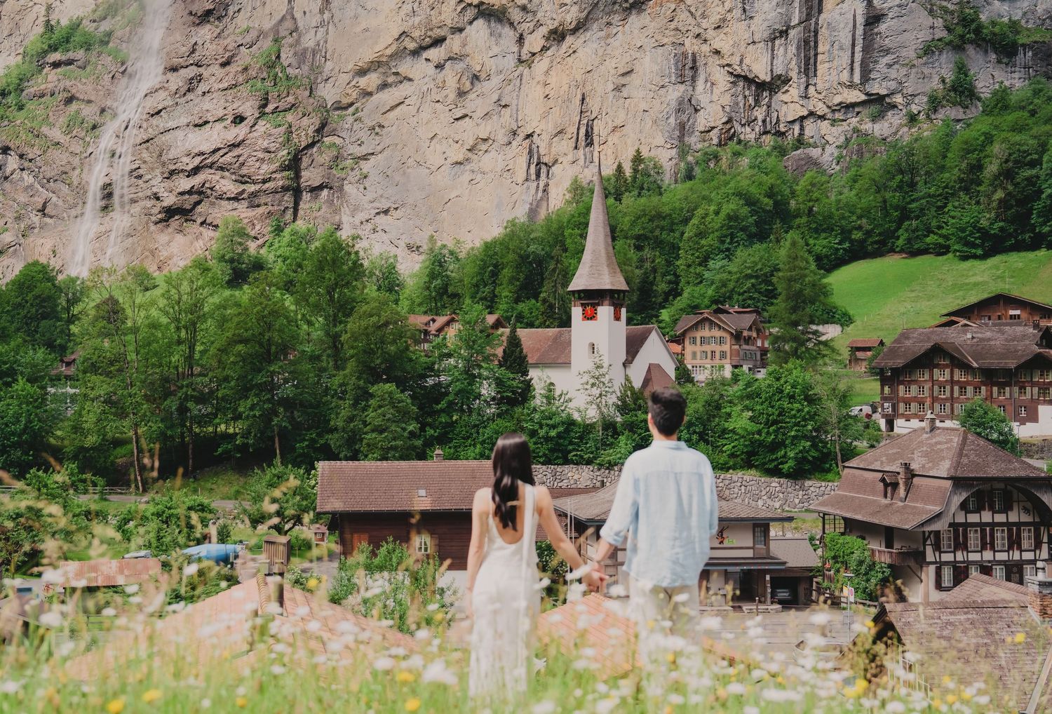 Couple holding hands overlooking charming Swiss village of Lauterbrunnen with waterfall and church steeple amid lush greenery.