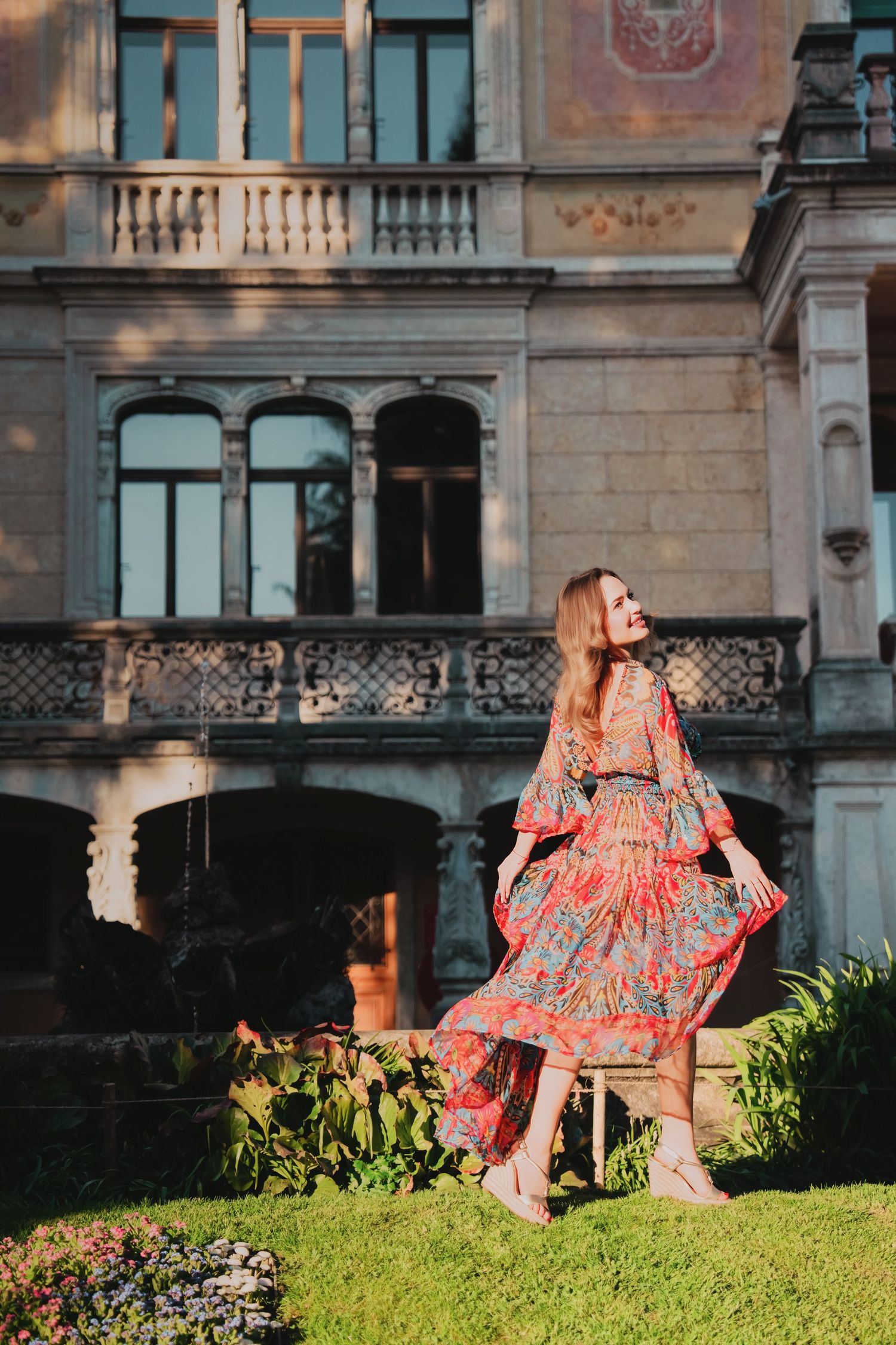 Elegant woman in vibrant boho maxi dress posing before ornate historic European building facade.