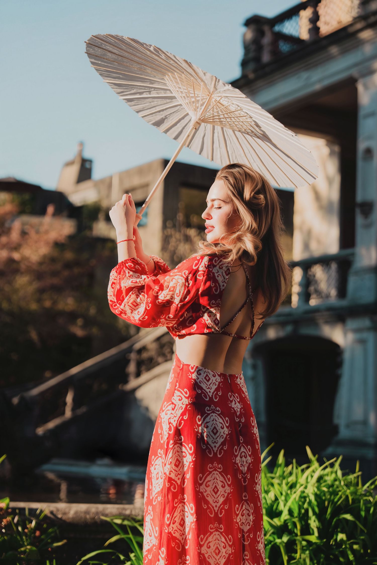 Blonde woman in red patterned crop set holding white parasol in sunlit garden near stone building.