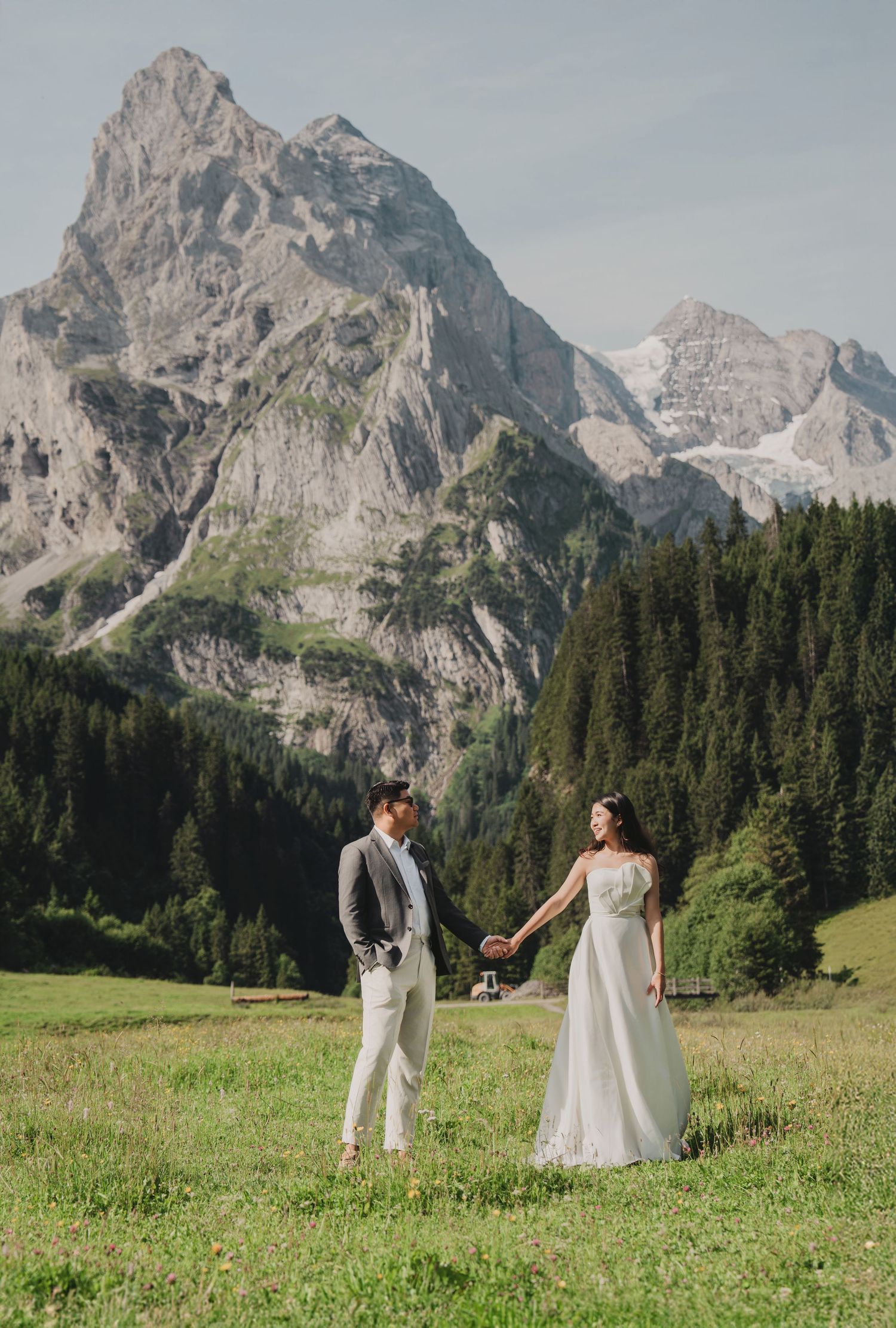 Bride and groom hold hands facing each other in sunlit Swiss alpine meadow with mountain backdrop.