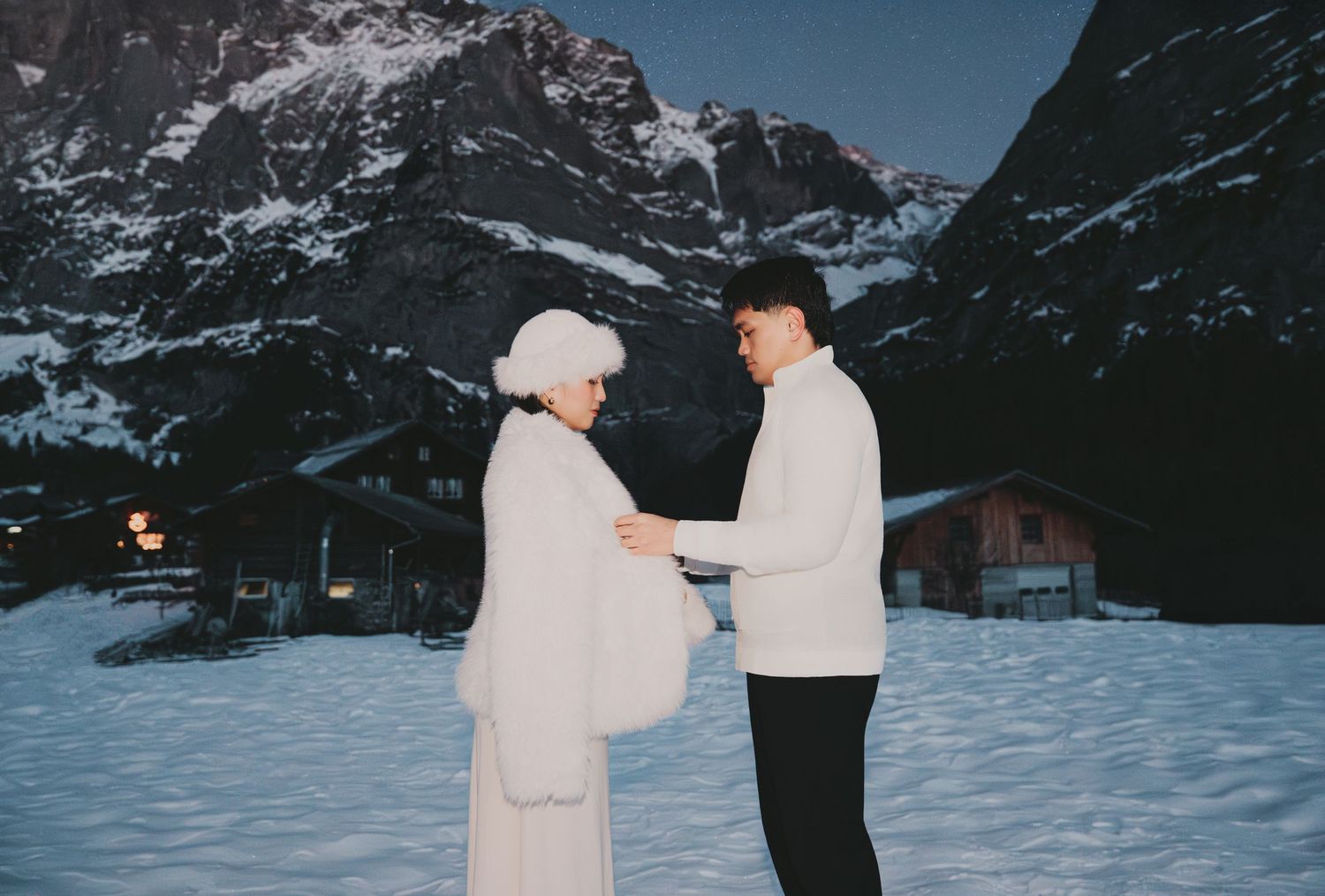 Couple holding hands facing each other in vintage-toned photo with snowy mountain village backdrop.