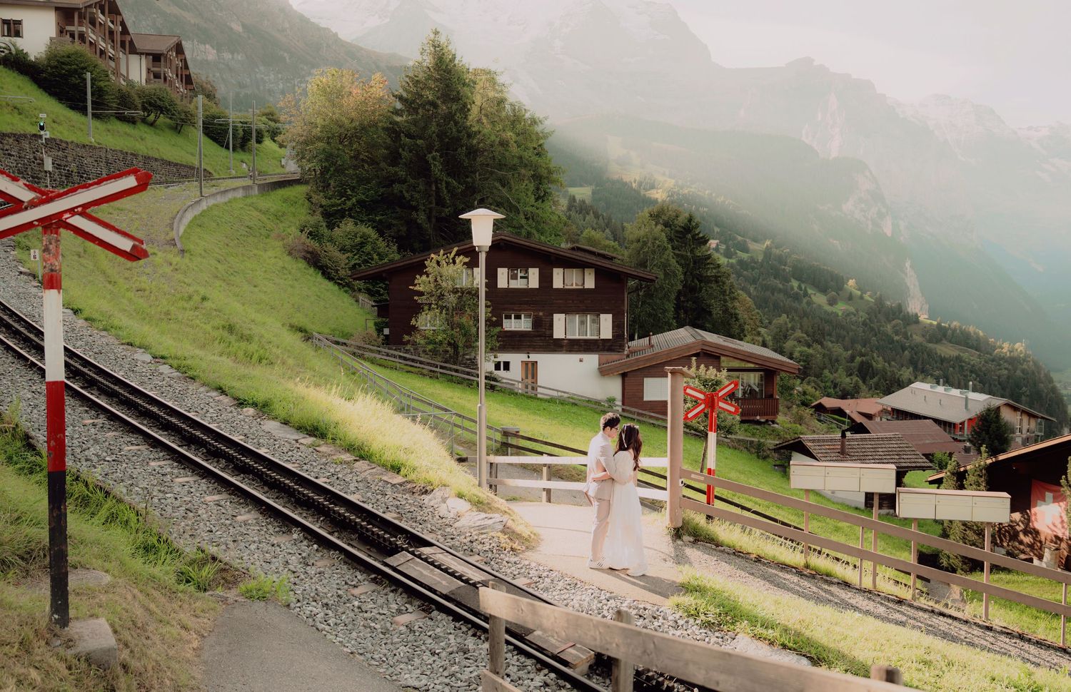 Bride in white dress walks along a scenic Swiss mountain railway track with alpine village and misty peaks backdrop.