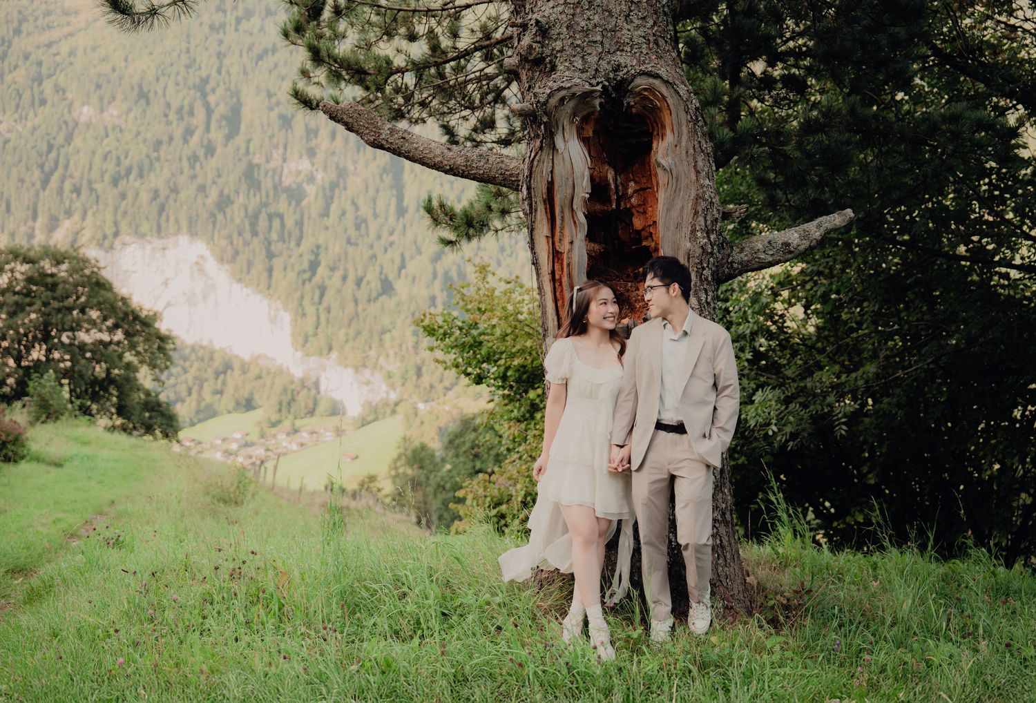 Smiling wedding couple stands beside a hollow ancient tree on a green alpine meadow with mountain valley backdrop.