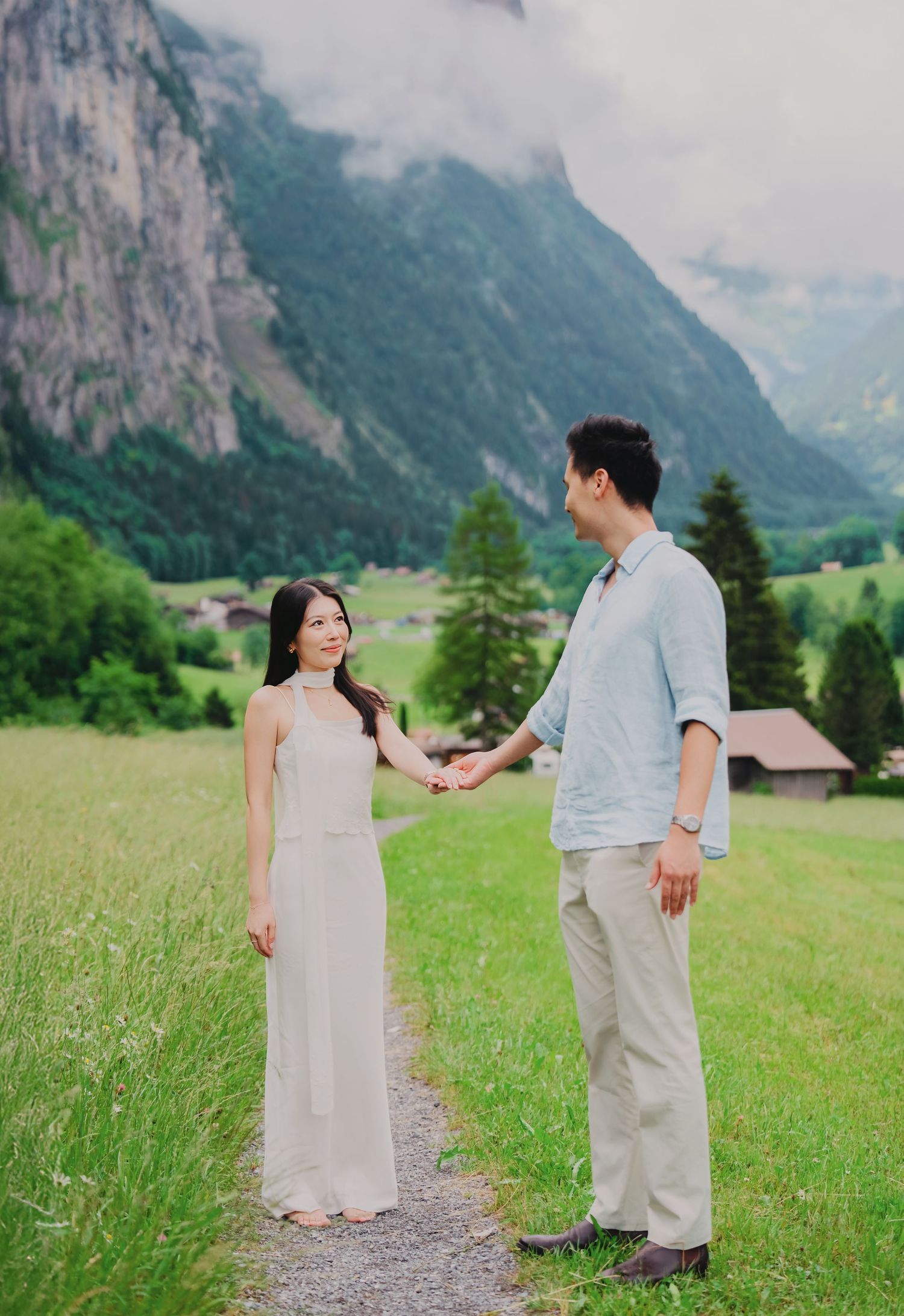 Couple holding hands on a scenic alpine meadow path with misty mountains in the background.