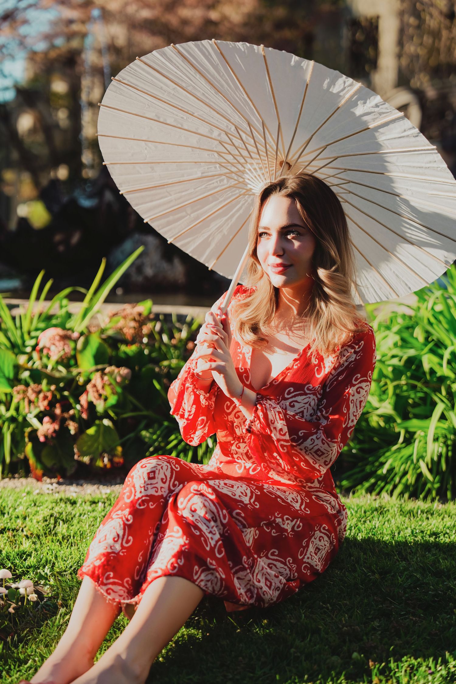 Blonde woman in red floral dress sitting on lawn with white paper parasol umbrella.