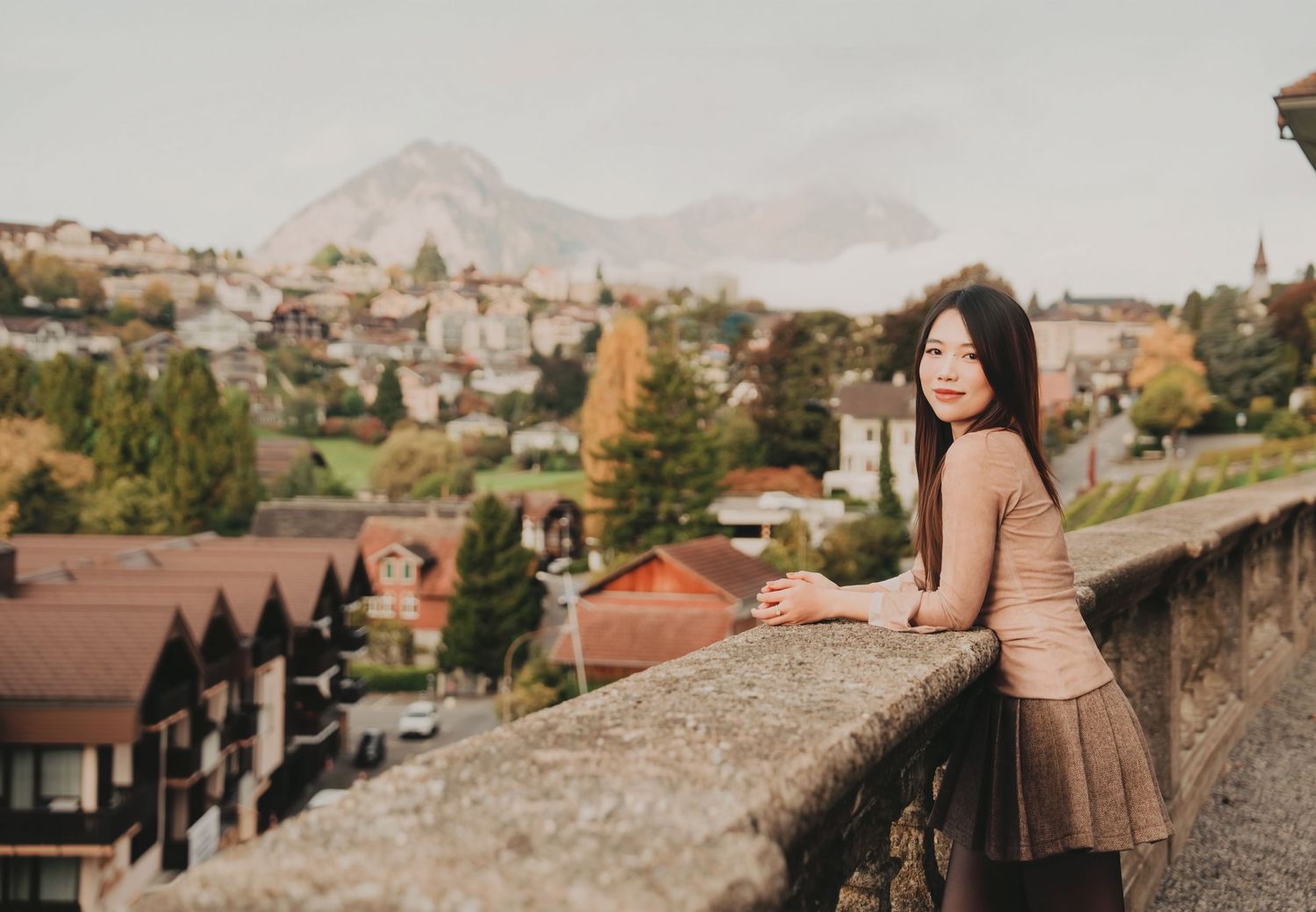 Young woman leaning on a stone wall overlooking a European mountain town with autumn trees.