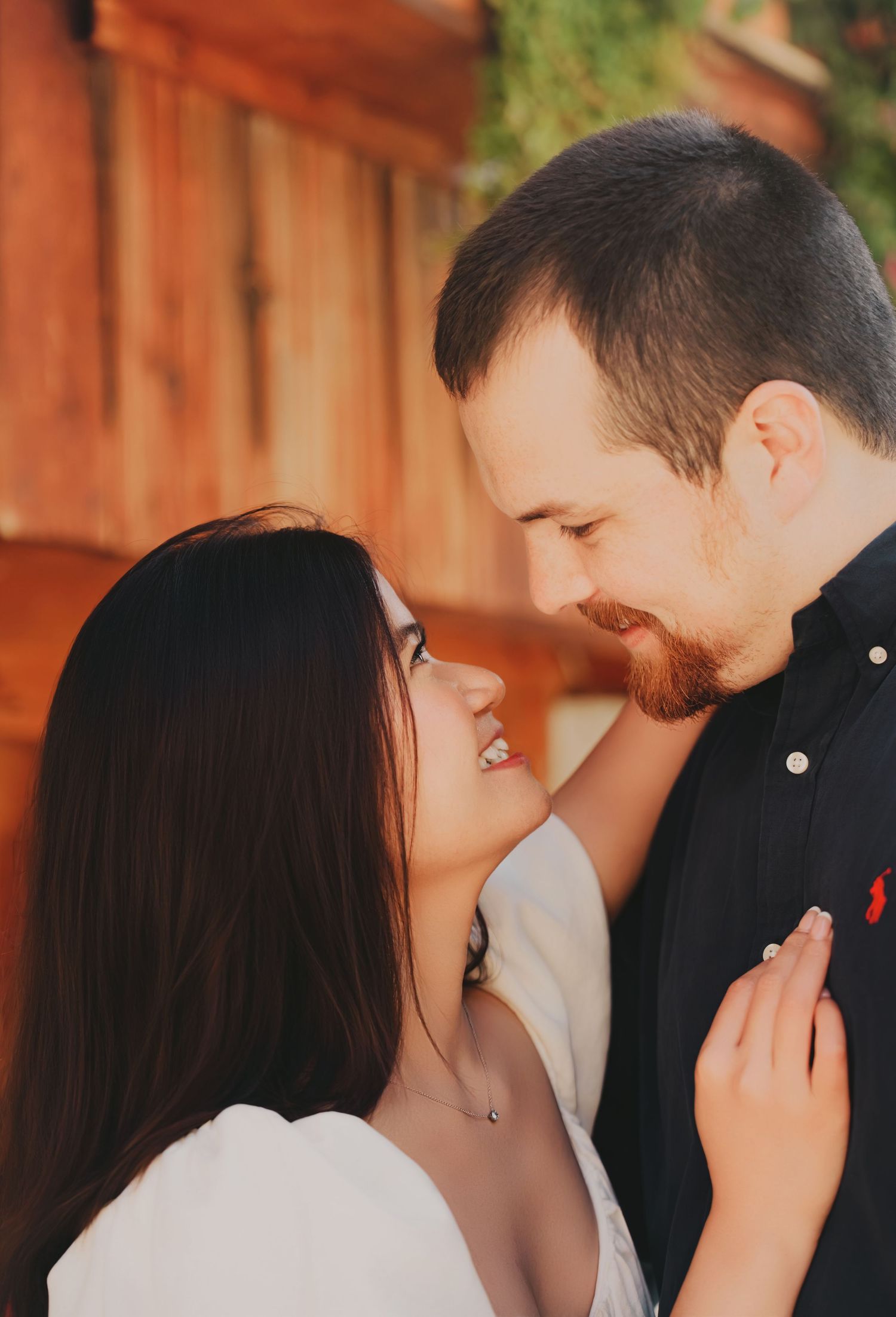 A couple sharing a romantic moment, smiling at each other near a wooden wall in warm golden light.
