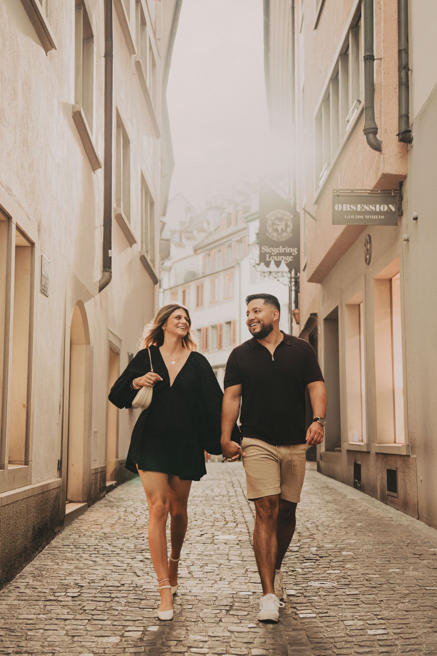 Happy couple holding hands walking down a sunlit cobblestone alley in Zurich.