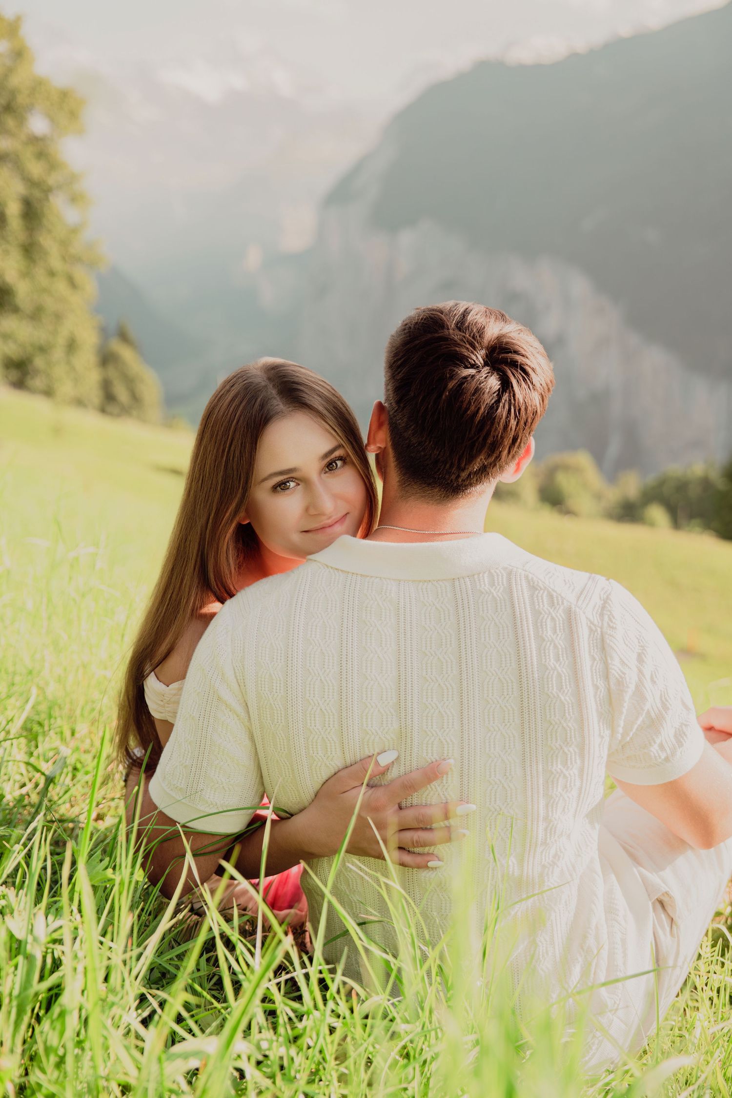 A woman hugs her partner from behind while sitting in a green meadow with mountain views.