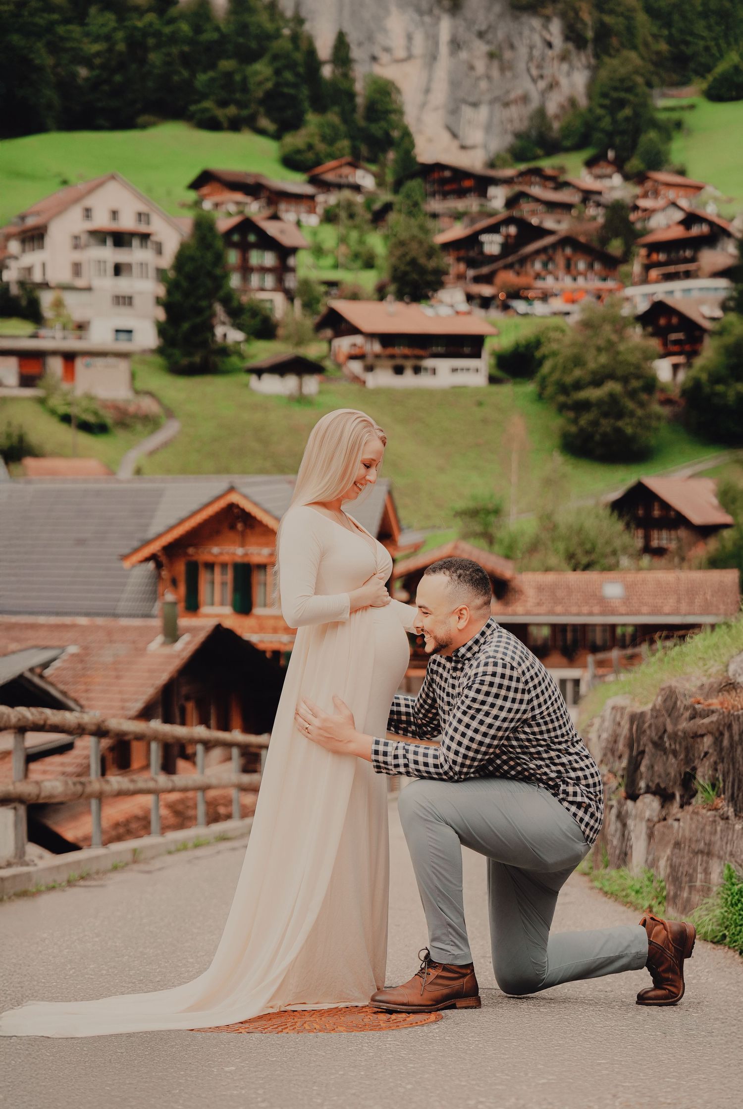 Man kneeling to kiss pregnant partners belly on bridge in scenic Lauterbrunnen Switzerland village setting.