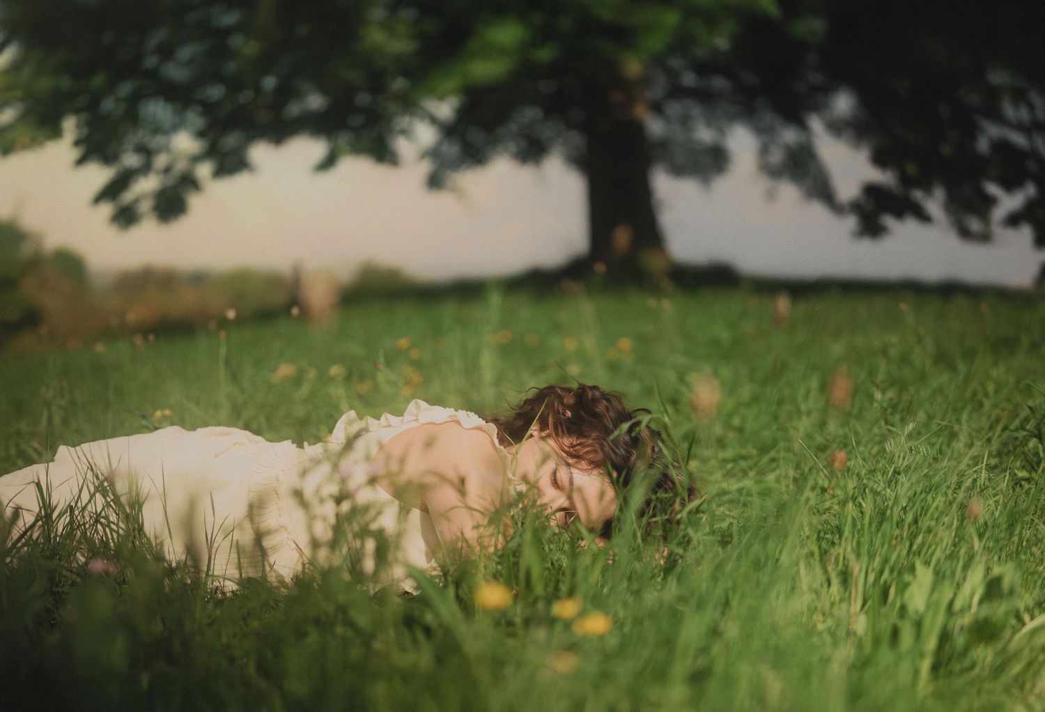 Woman in white dress lying face down in green grass field with yellow wildflowers and tree.