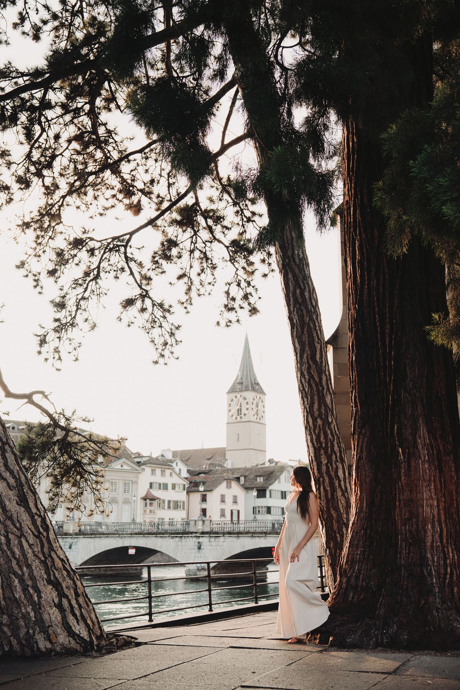Woman in white dress leaning against large tree by Zurich river with historic church spire backdrop.