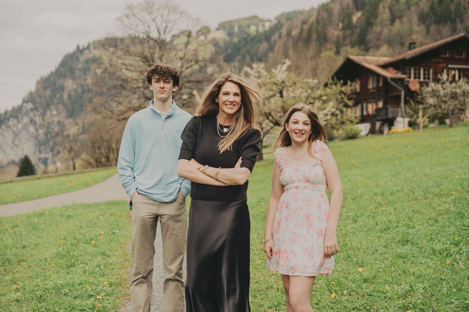 Happy family of three laughing together on grassy hillside with Swiss mountain chalet backdrop.