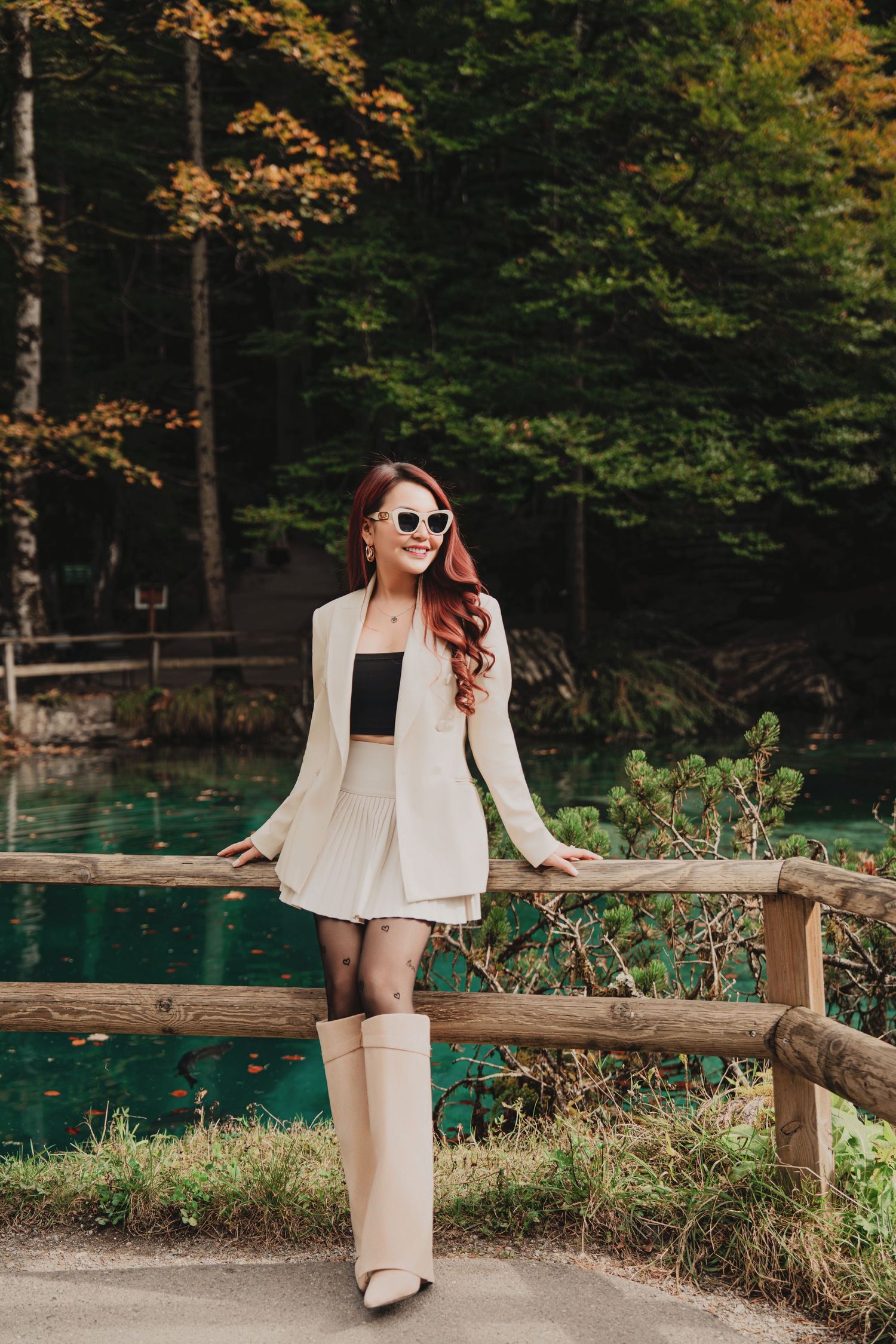 Stylish woman in white blazer and skirt set with sunglasses posing by turquoise forest lake on wooden fence.