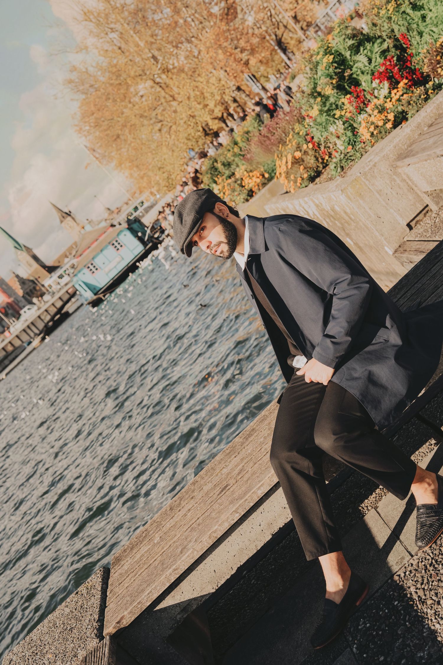 Fashionable bearded man in dark coat and flat cap poses on waterfront steps with autumn foliage.