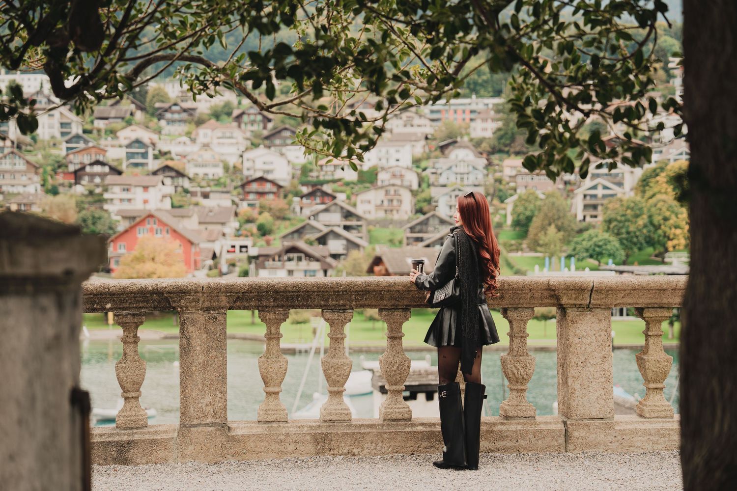 Red-haired woman in black outfit stands at stone balustrade terrace overlooking picturesque Swiss alpine village and lake.