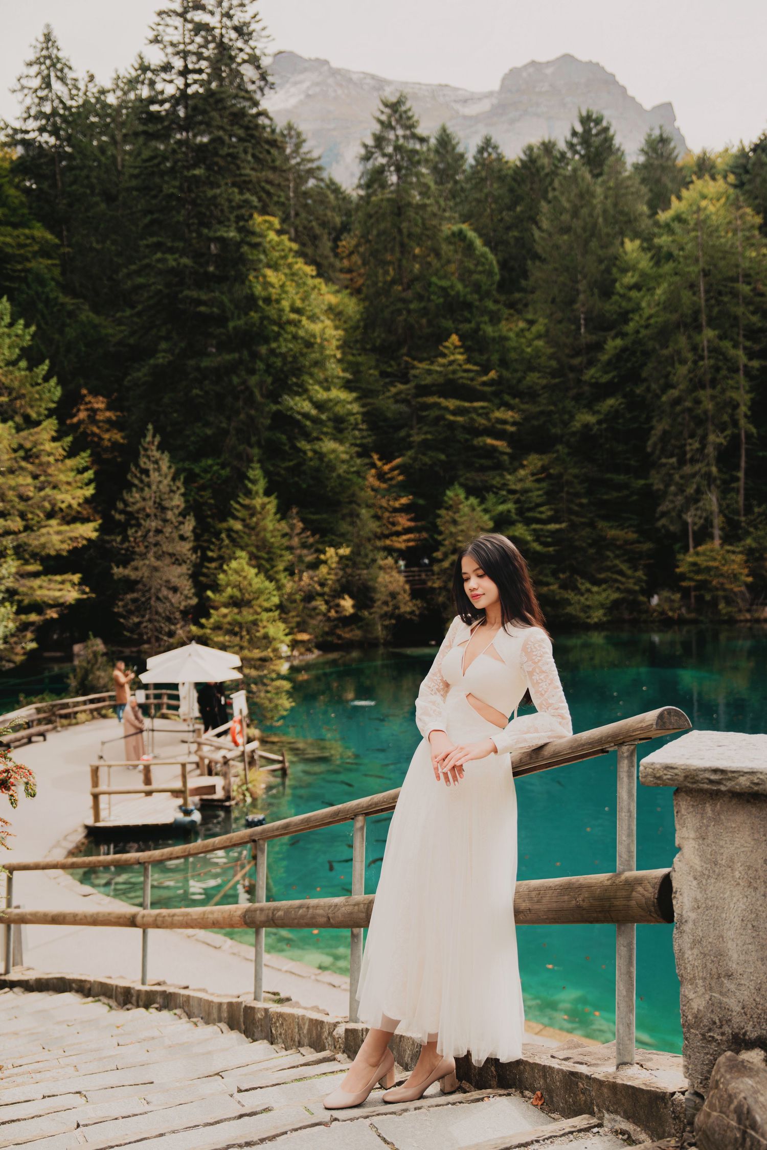 Elegant woman in white dress stands by railing overlooking stunning turquoise alpine lake.