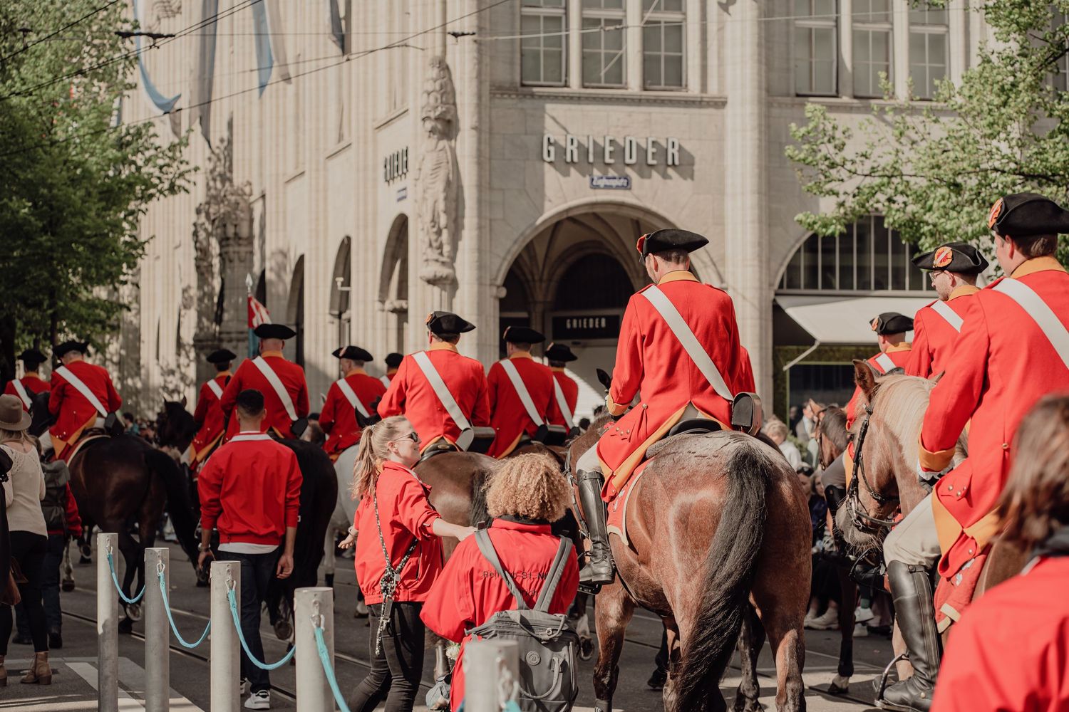 Mounted guards in red historical uniforms parade through a city street past spectators.