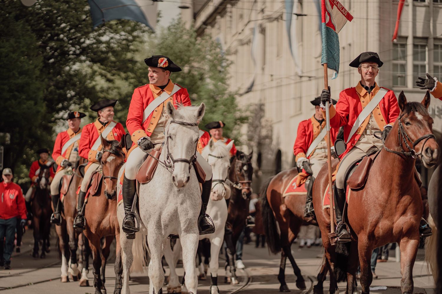 Group of riders in red historical uniforms on horseback parade through a European city street.