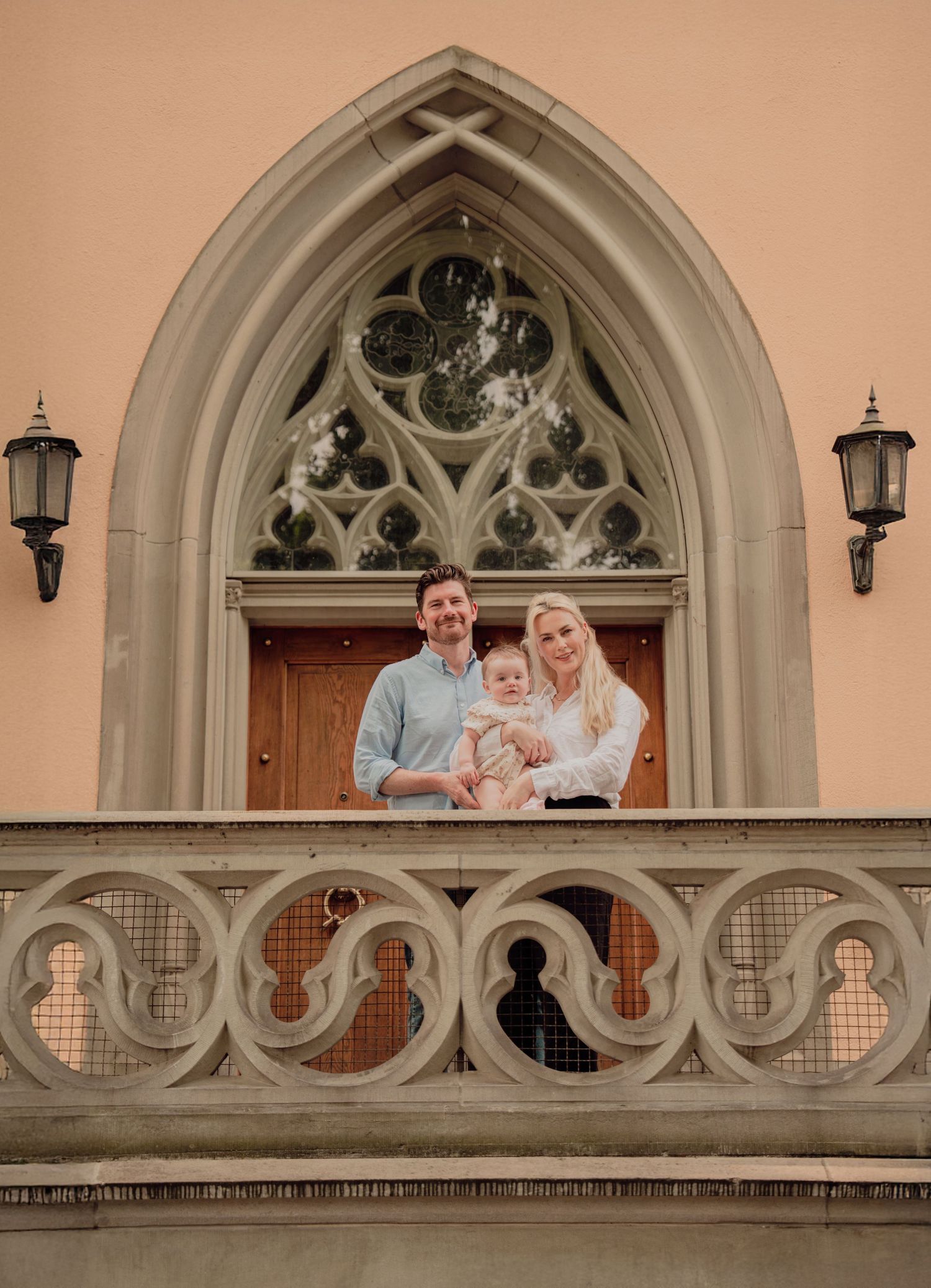 Family of three posing on ornate Gothic church balcony with arched window backdrop.