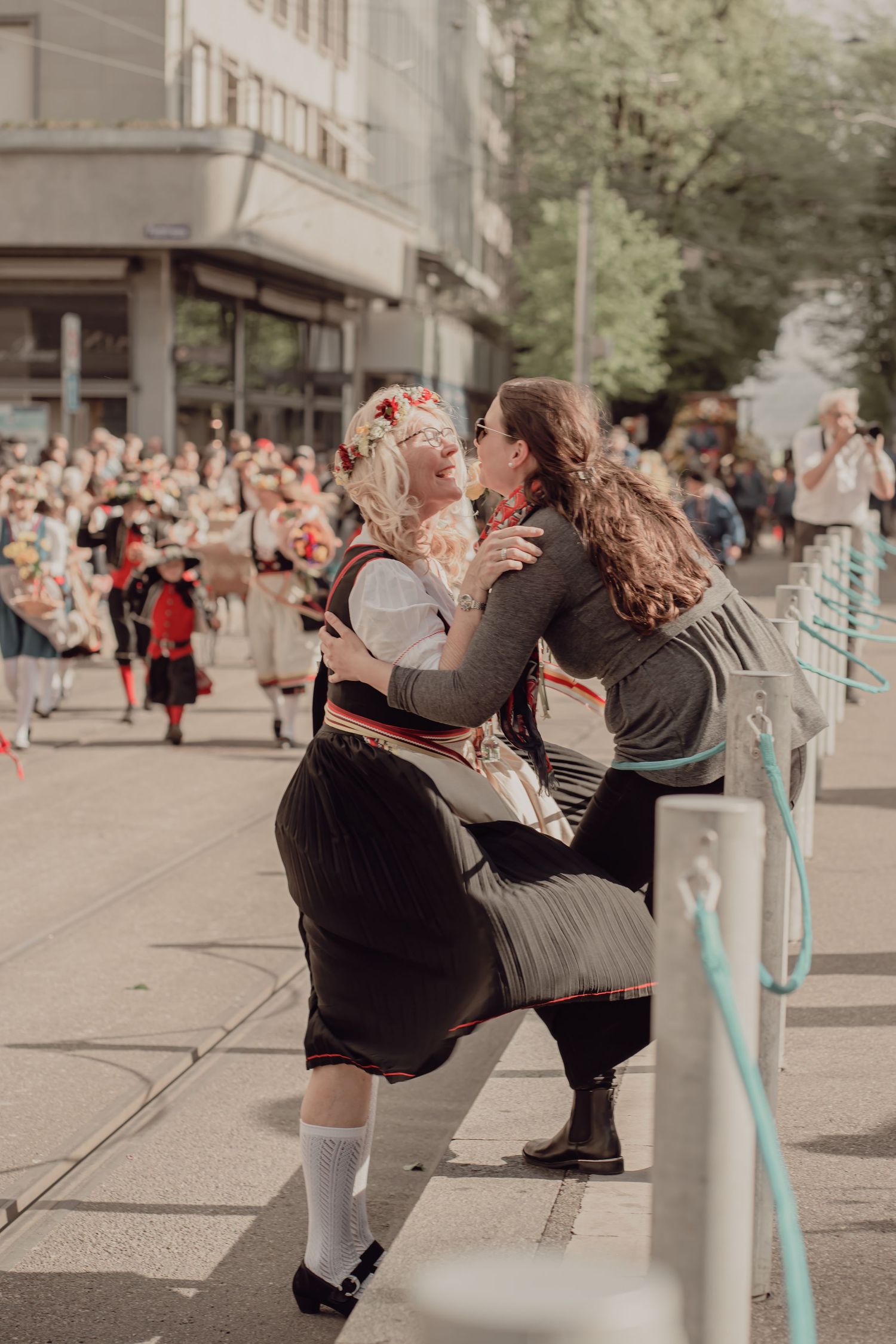 Two women in traditional folk costumes share a kiss during a cultural parade on a city street.