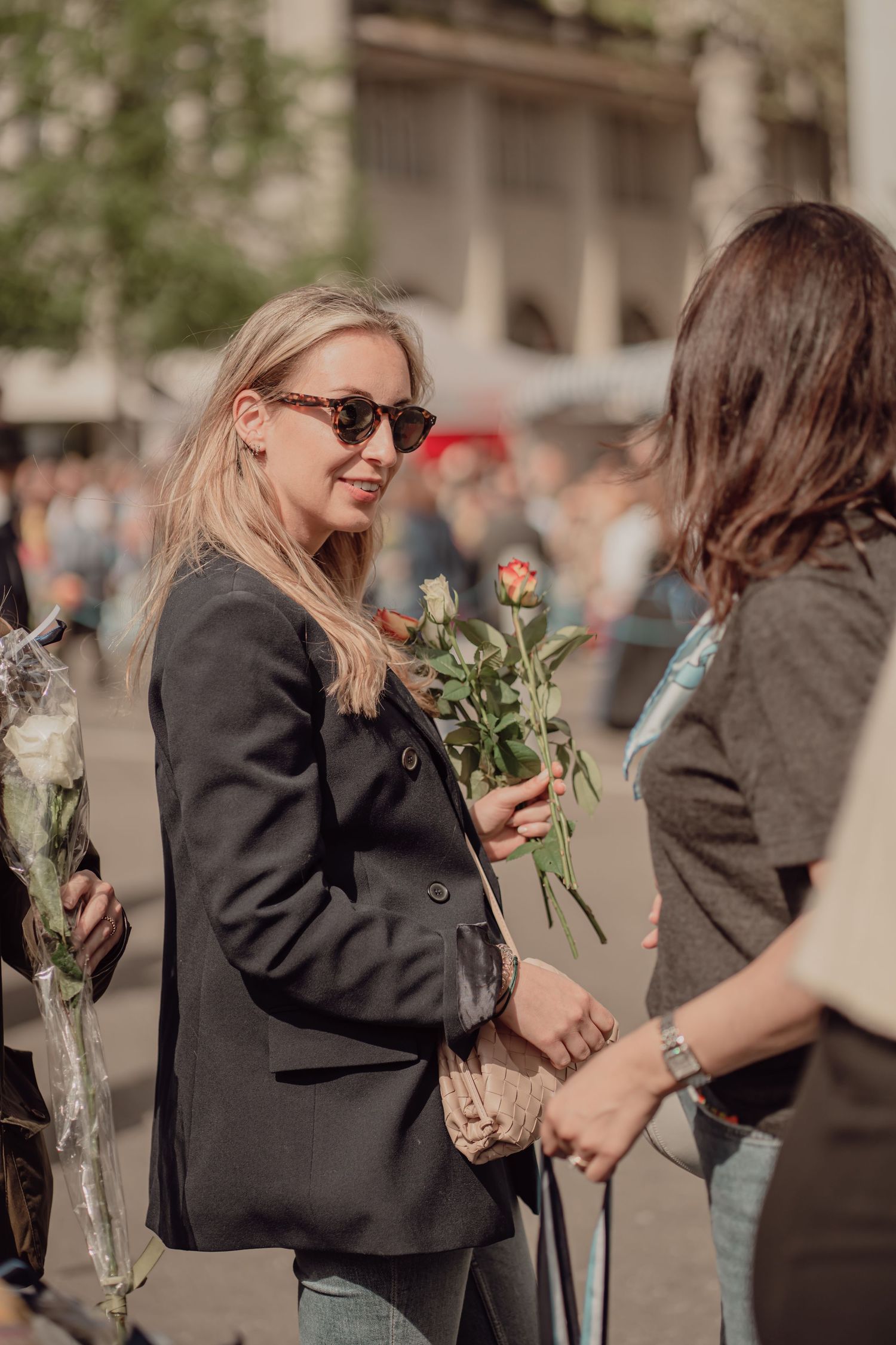 Smiling blonde woman in sunglasses holds roses at a lively outdoor public event.