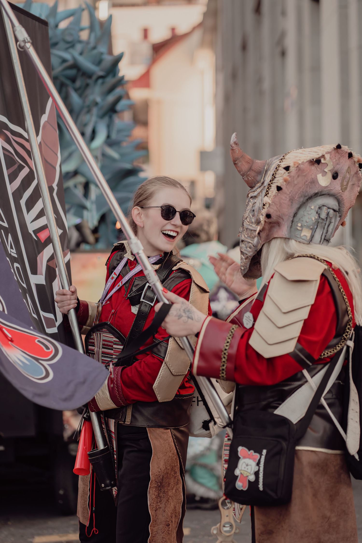 Two cosplayers in matching red fantasy armor laugh together at an outdoor convention event.