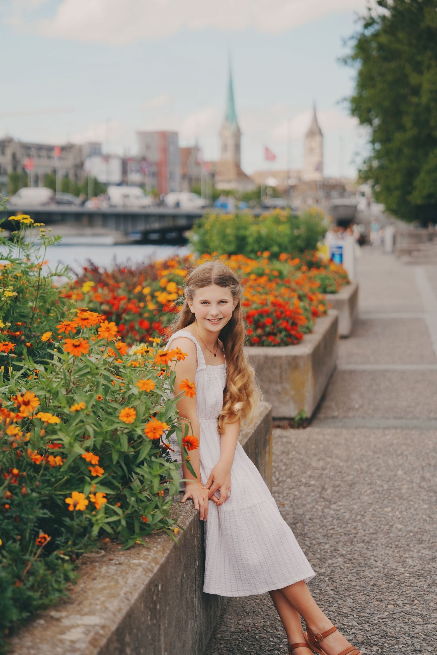 Smiling girl in white dress posing beside vibrant orange blooms with Zurich cityscape in background.