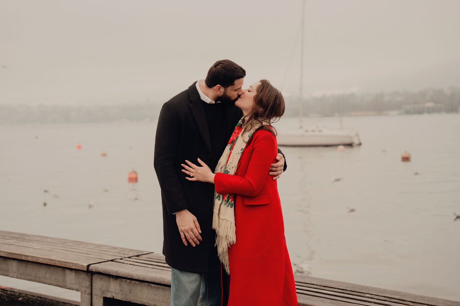 Couple sharing a kiss by a misty lake, woman in red coat and floral scarf embracing man in black.