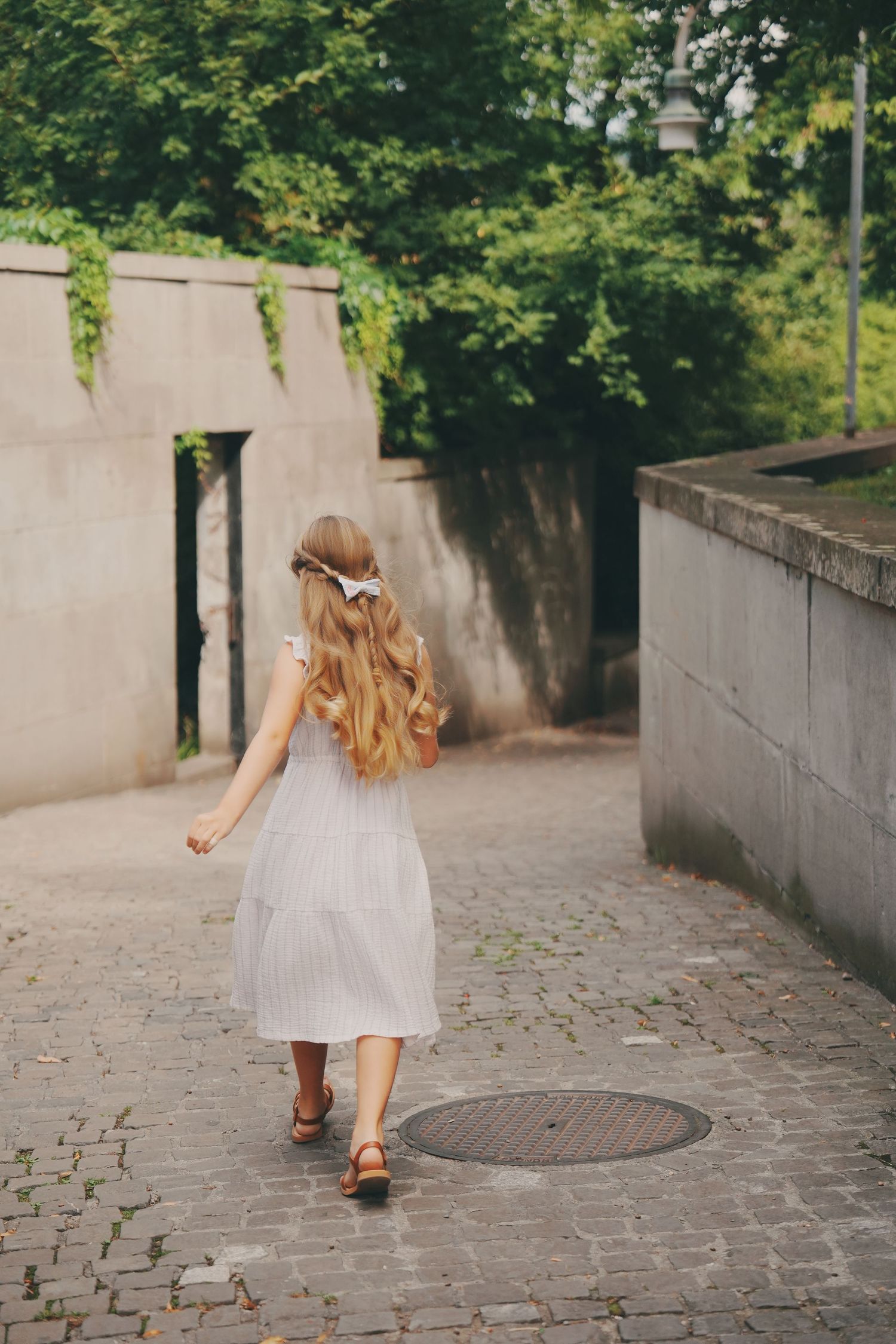 Young girl in white dress with hair bow walking along a cobblestone path.