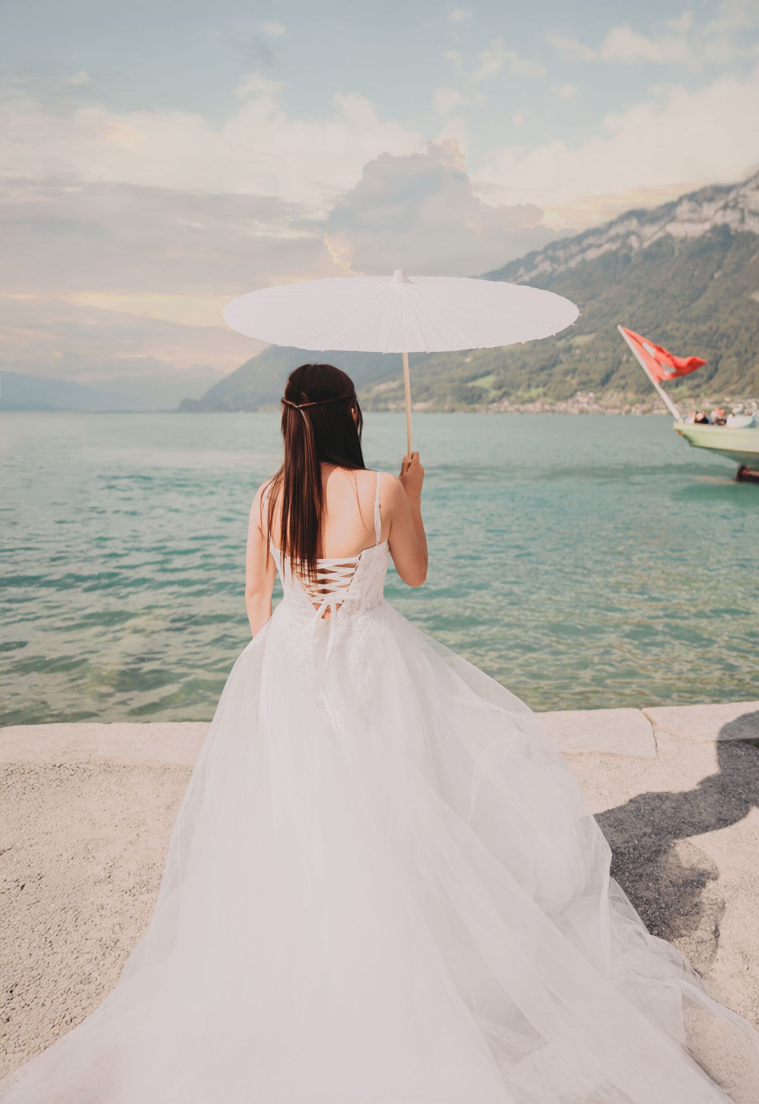 Bride in white wedding gown holding parasol by turquoise Swiss lake with mountain backdrop.