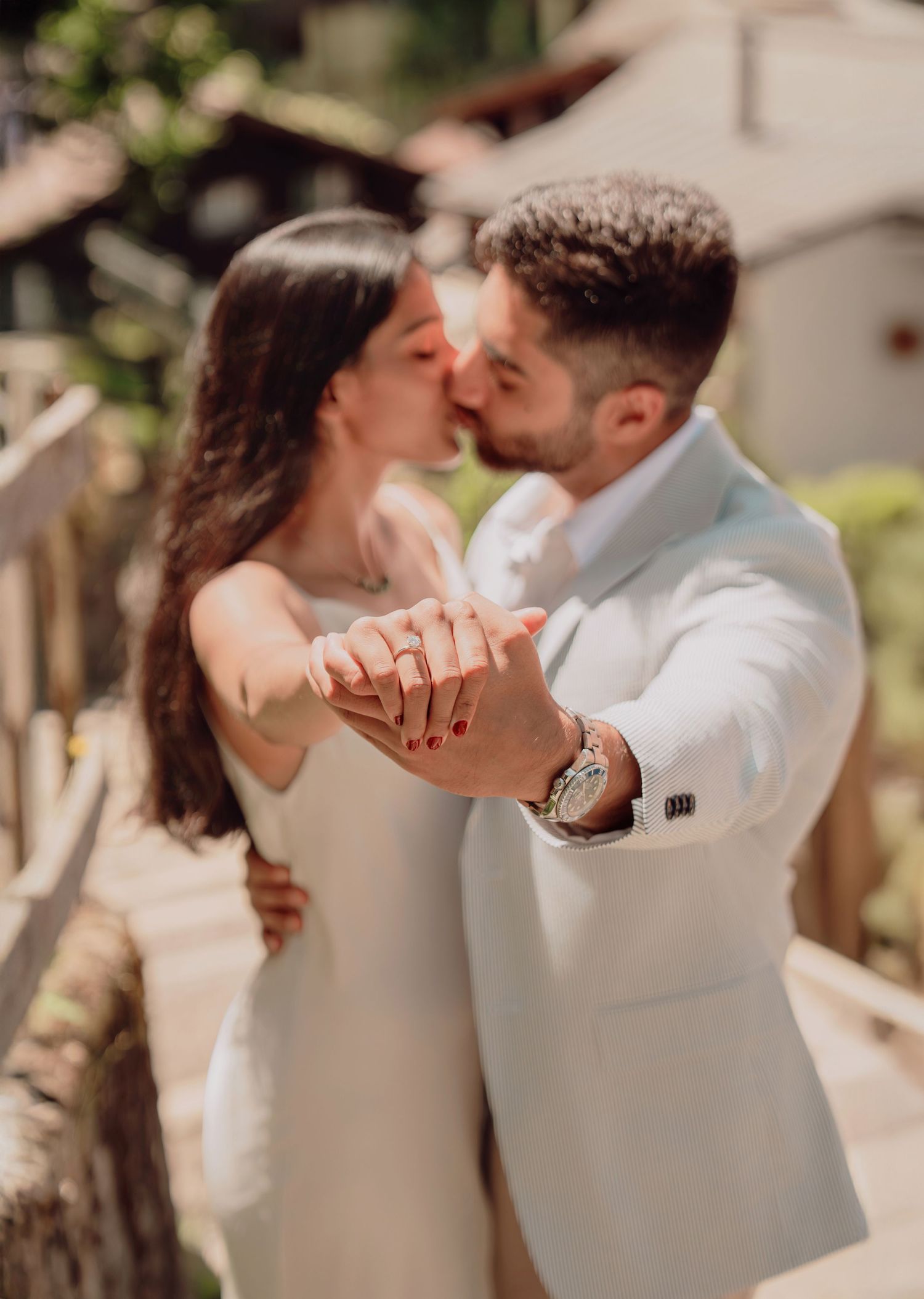 Couple in white outfits sharing a kiss while holding hands during an outdoor engagement photo session.