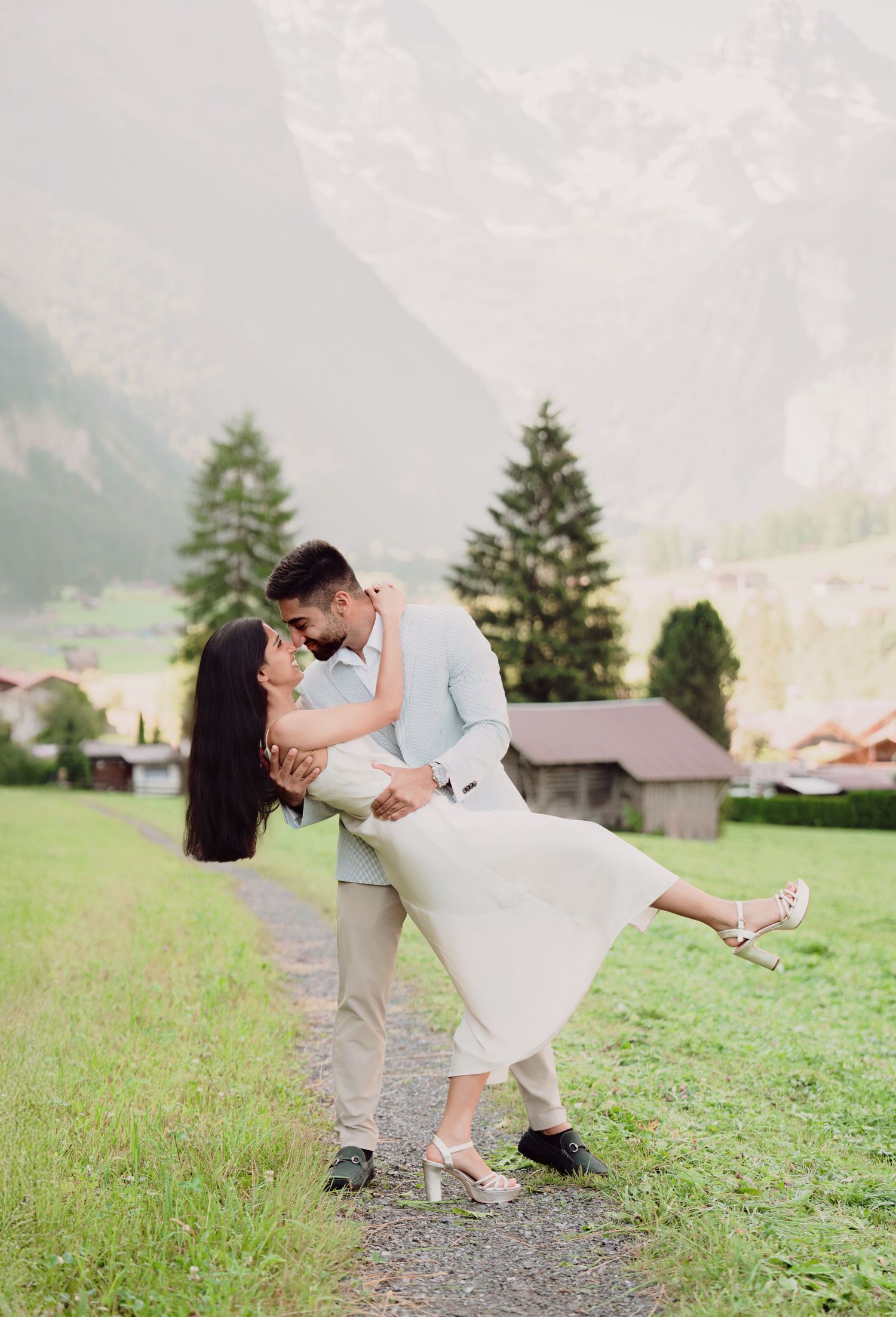 Romantic couple sharing a dip kiss on a path with misty Swiss mountain scenery behind.