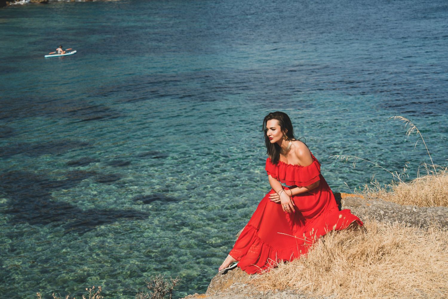 Elegant woman in flowing red dress relaxing on coastal rocks above clear blue water.