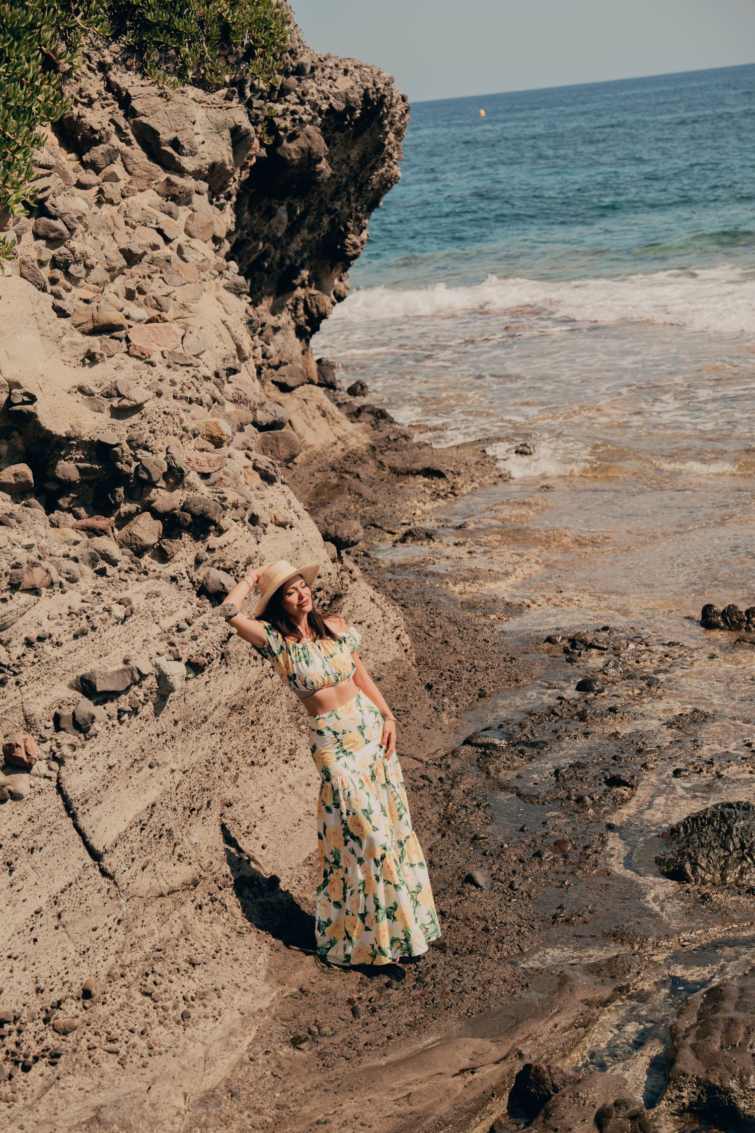 Woman in floral crop top and maxi skirt with straw hat posing on rocky beach.