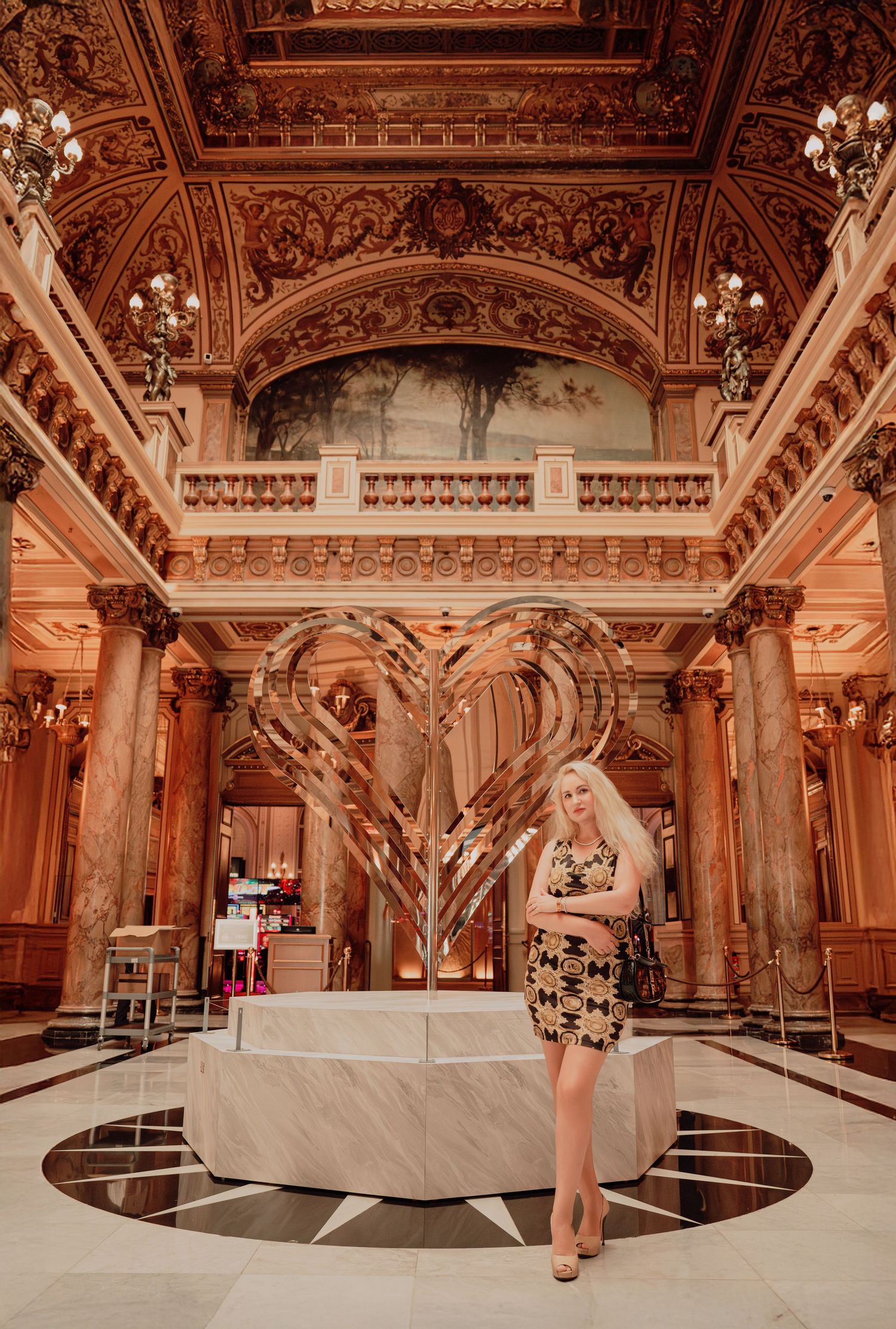 Blonde woman in patterned dress standing before a large golden heart sculpture in an opulent baroque interior.