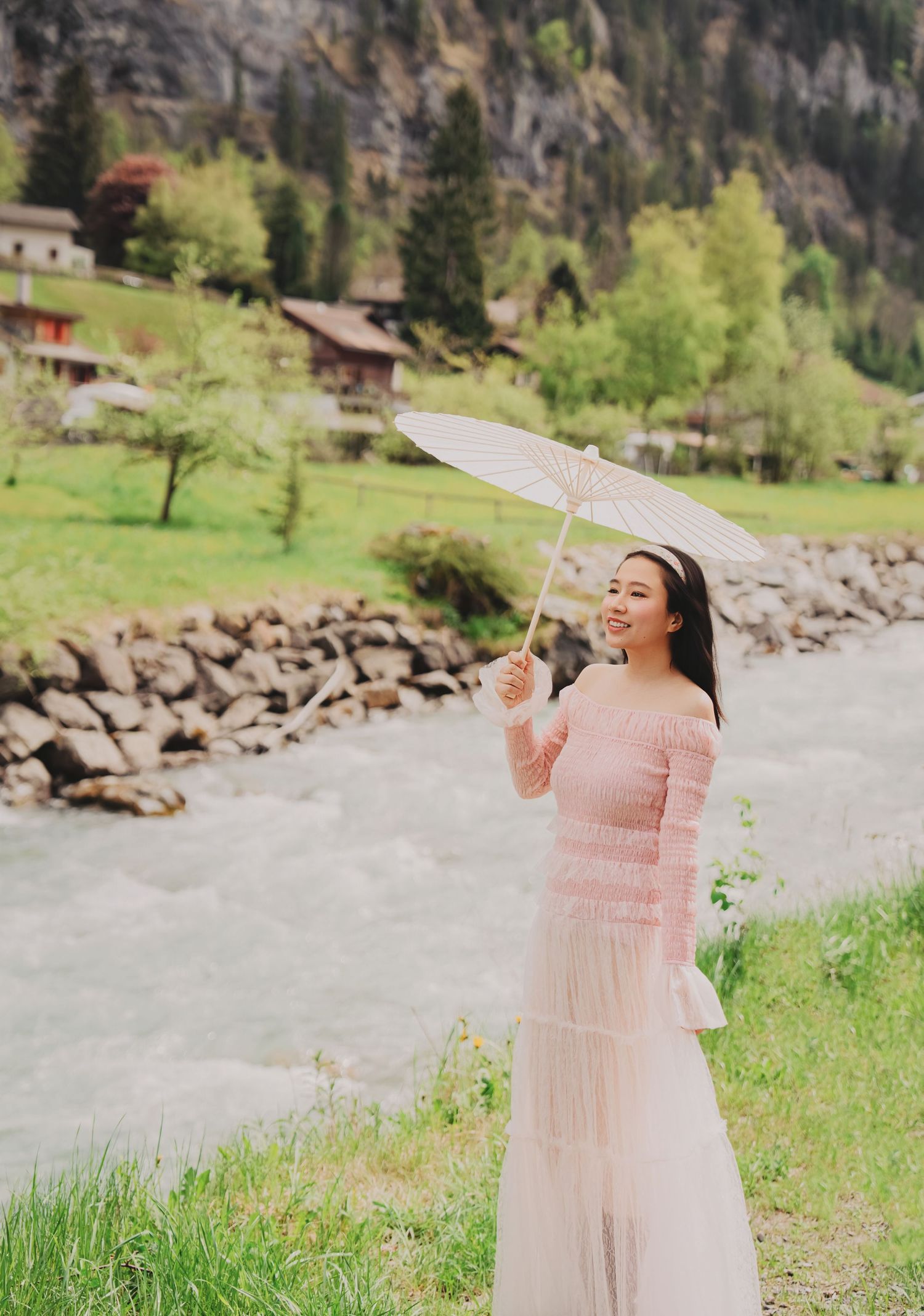 Woman in pink dress holding white parasol smiling by rocky river in Swiss alpine village.