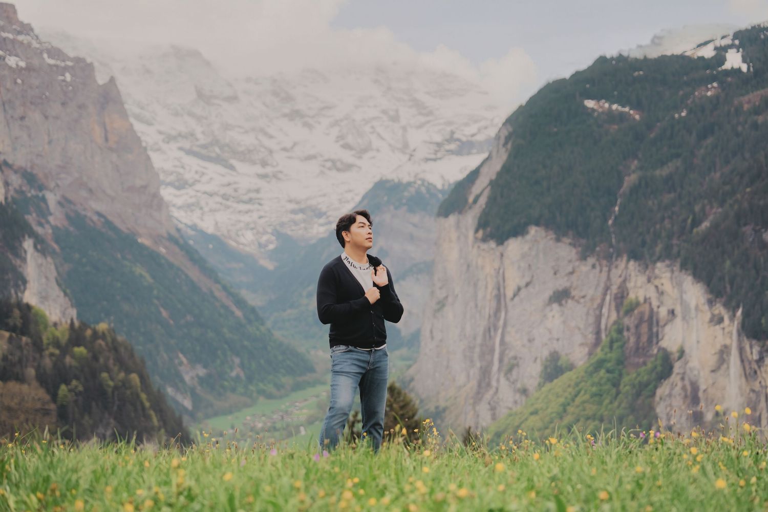 Man standing alone in a green meadow with dramatic snow-capped alpine mountains behind him.