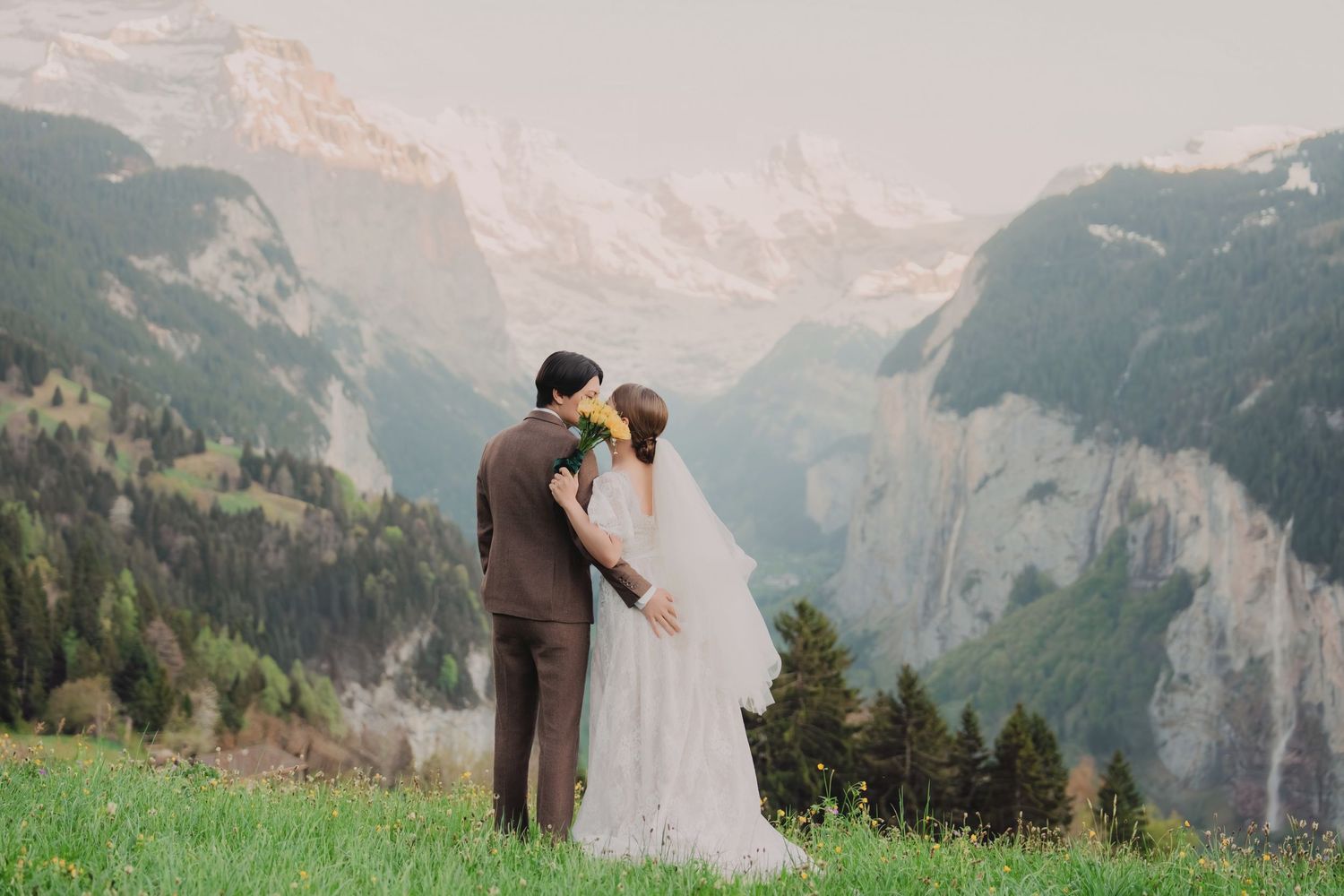 Wedding couple kissing on lush meadow with stunning Swiss mountain valley landscape behind.